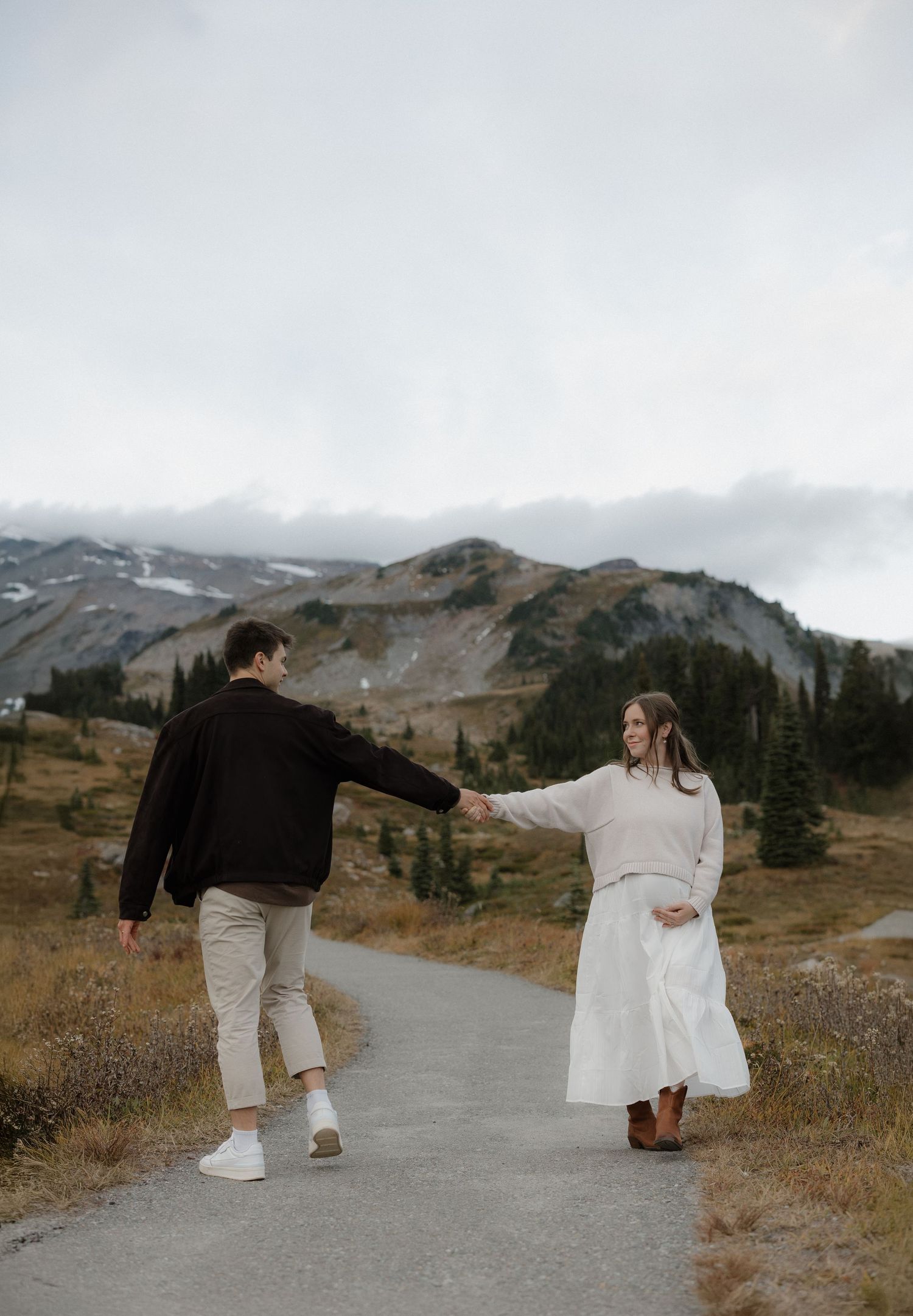 Couple holding hands on a mountain path, woman in white skirt and man in black jacket, with snowy peaks in the background.