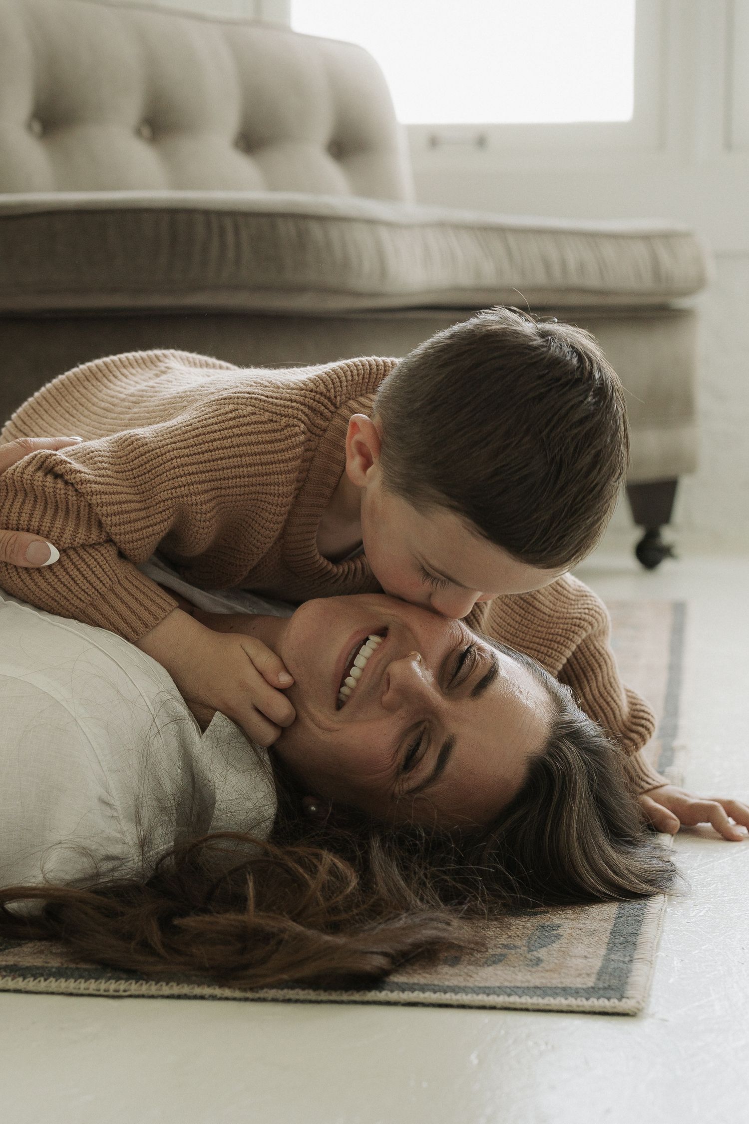 A joyful mother lying on a rug laughing as her young child leans over to give her a kiss on the cheek.