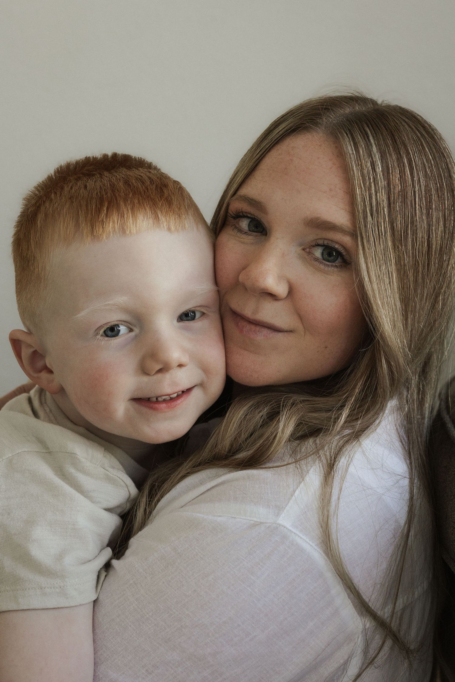 A smiling red-haired boy cuddles cheek-to-cheek with his blonde mother in a warm portrait photo.