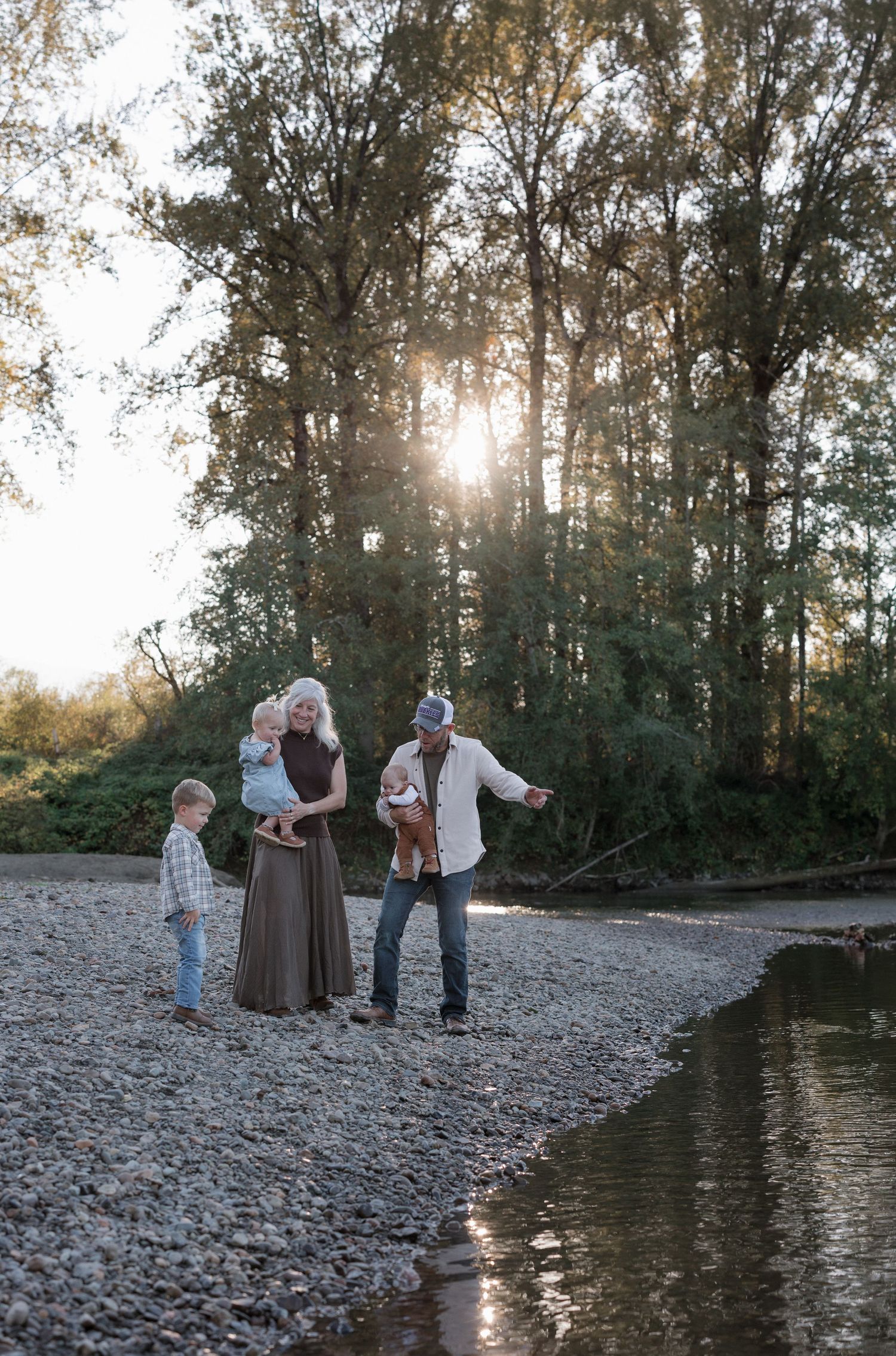 Family of four enjoying a sunny day by a rocky riverside surrounded by tall trees with golden sunlight filtering through.