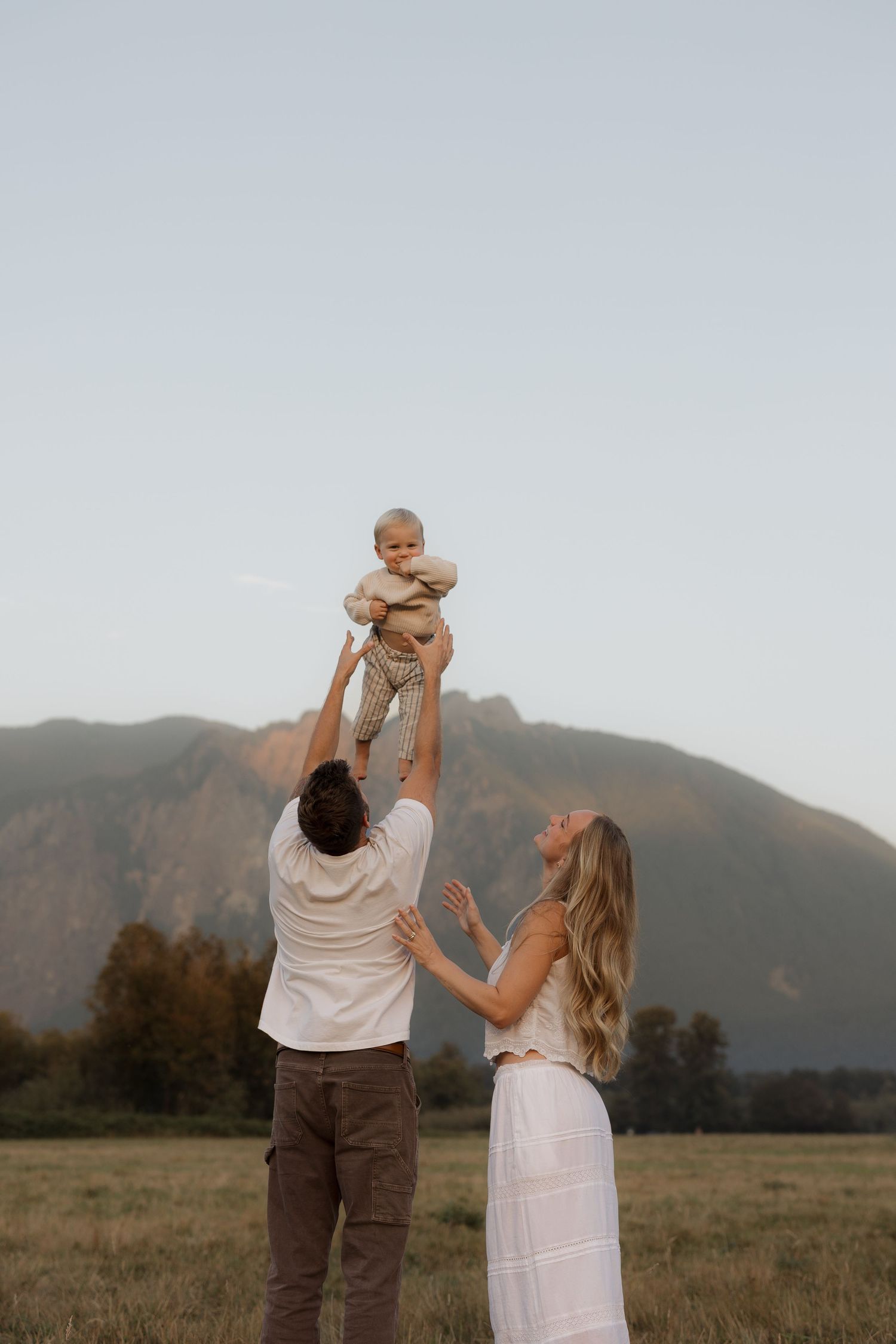 A joyful family in a mountain meadow, father tossing baby in the air while mother laughs, wearing neutral tones.