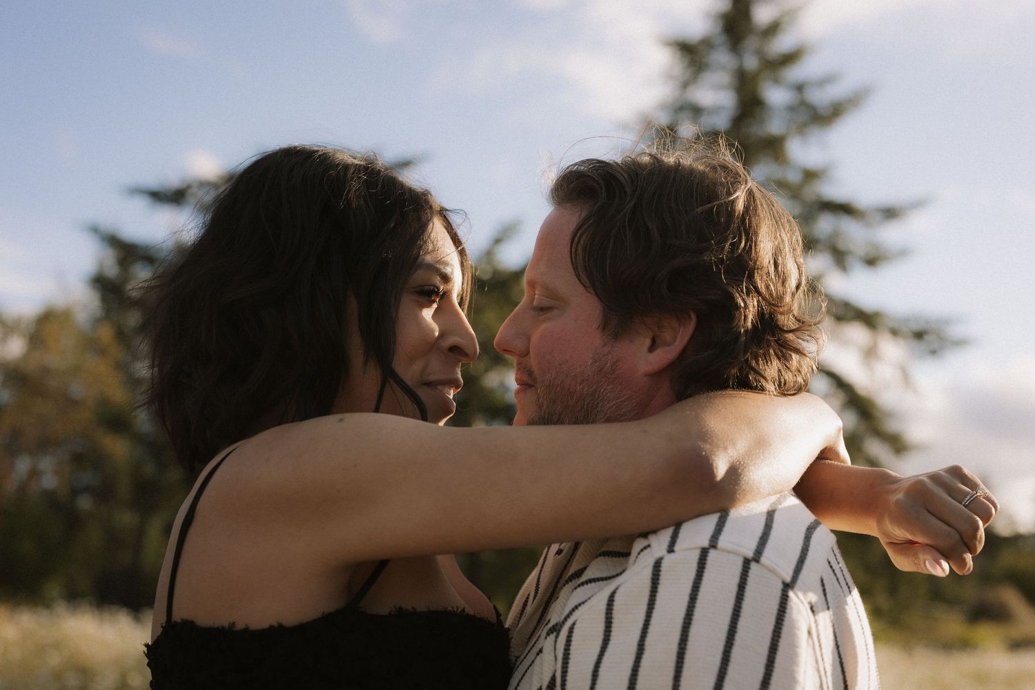 A couple embraces outdoors, gazing at each other with trees and blue sky in the background.