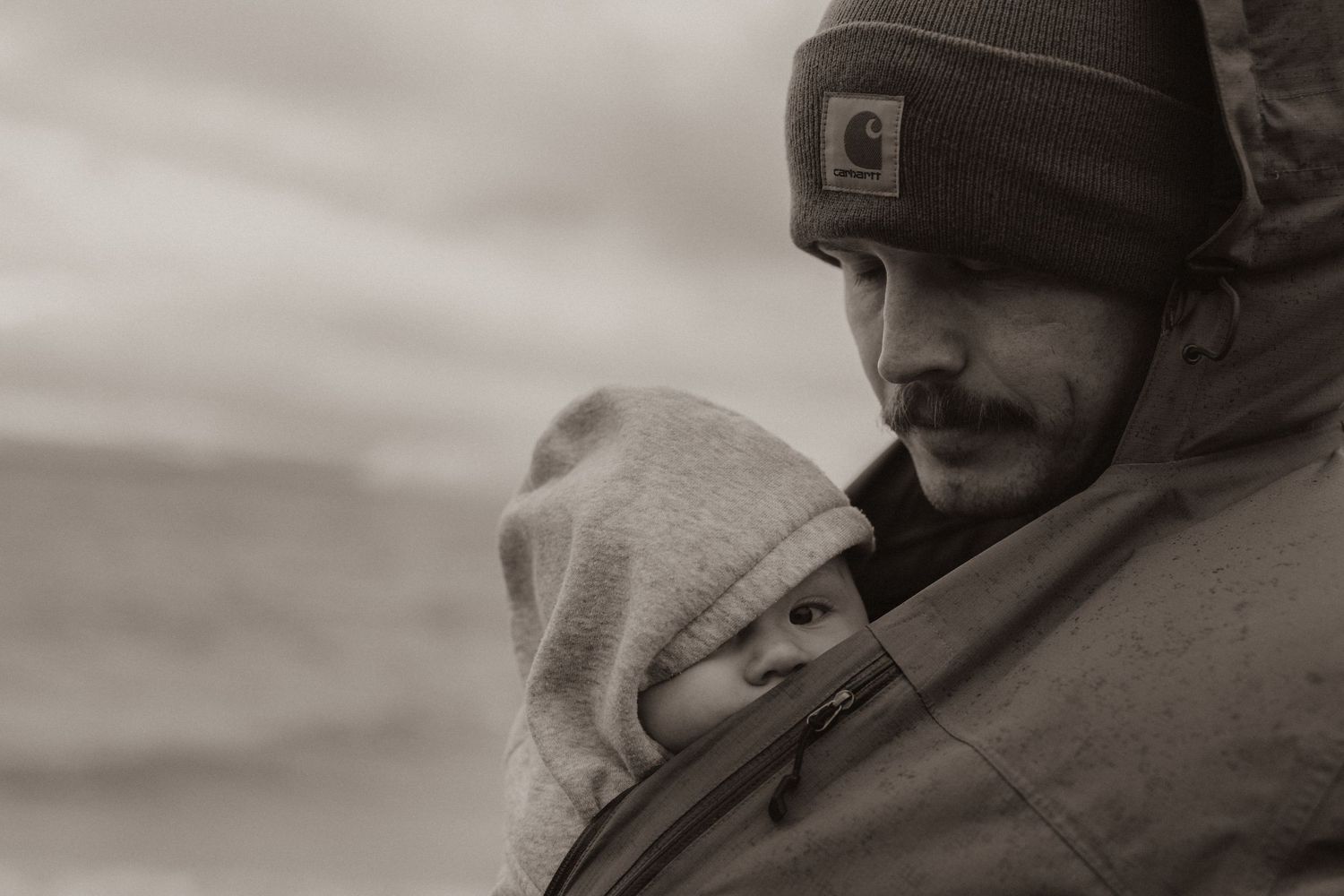 Black and white photo of a father with mustache and Carhartt beanie carrying a baby in a front carrier outdoors.