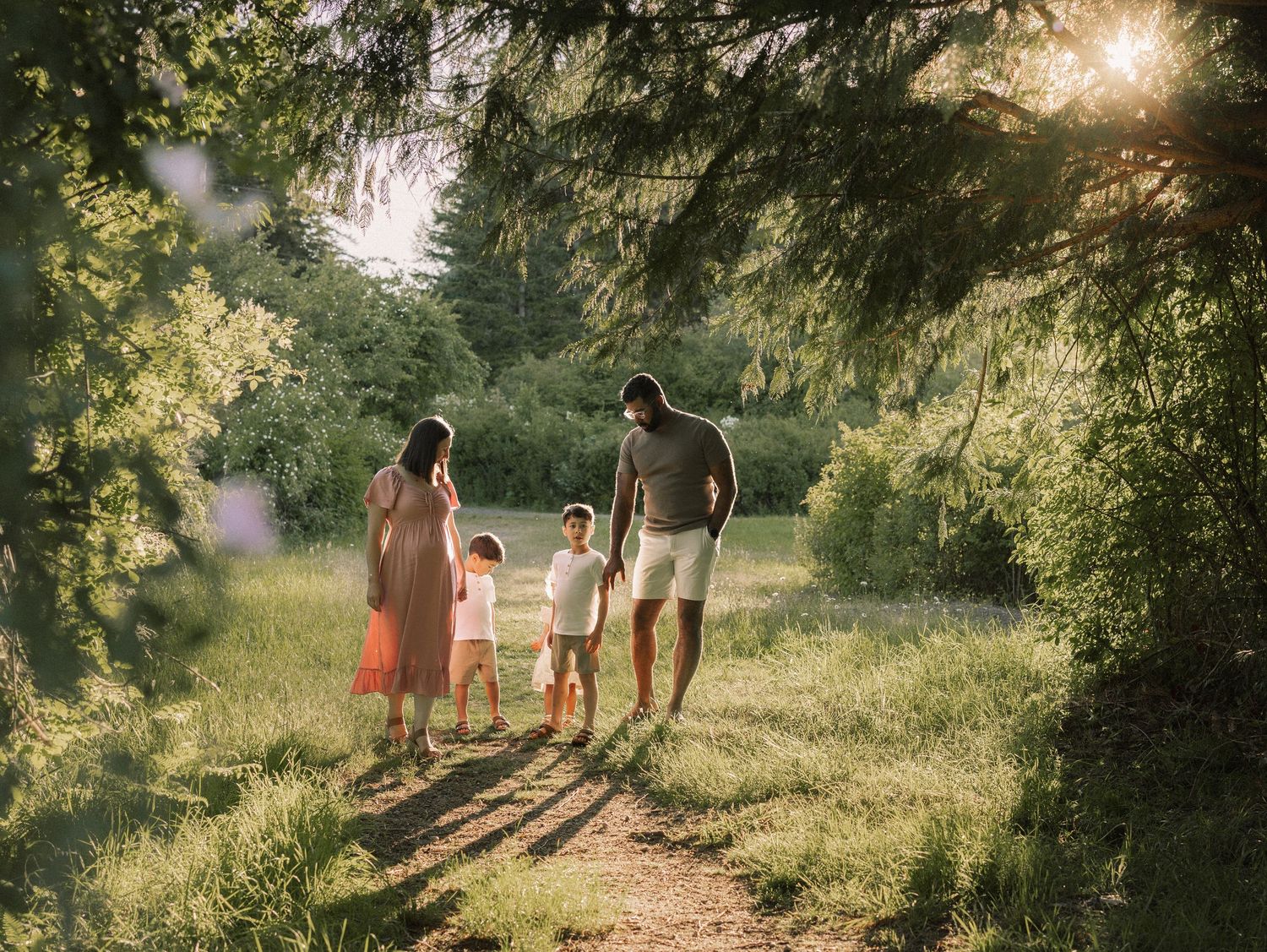 Family of four walking hand-in-hand through a sunlit forest path surrounded by lush green trees.