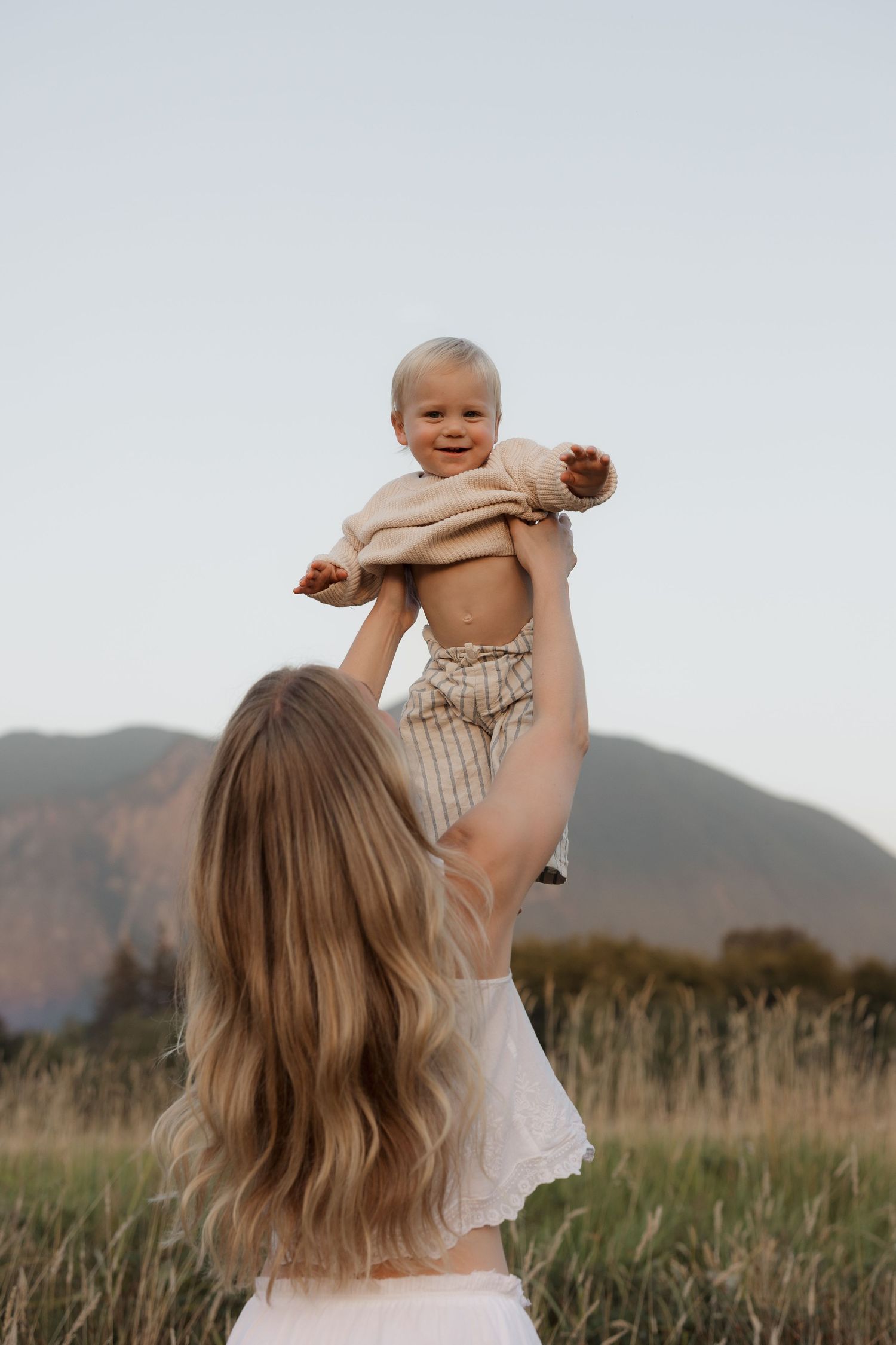 A mother lifts her smiling baby in the air outdoors in a field with mountains in the background.