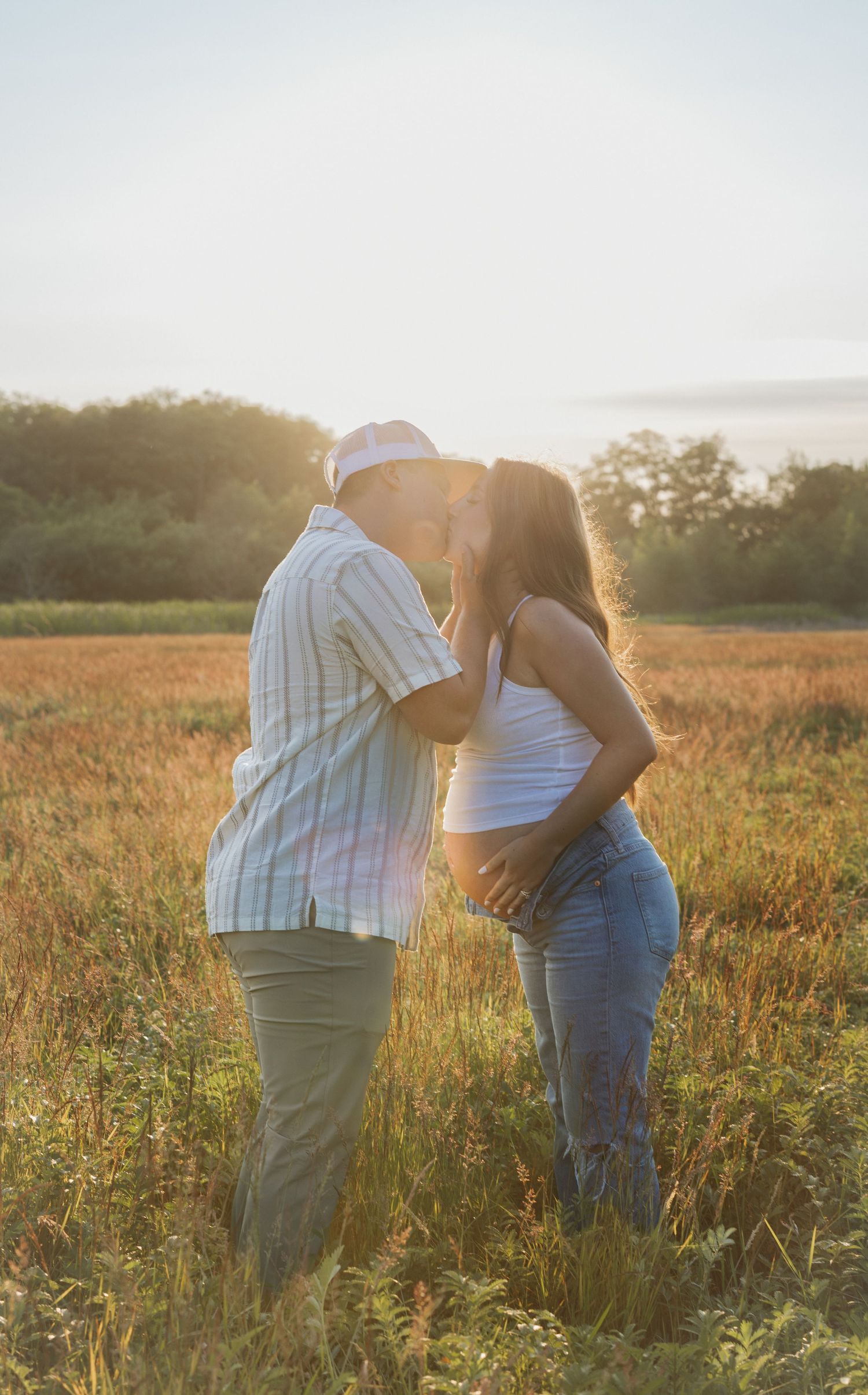 Couple sharing a kiss in a sunlit golden field during a warm summer maternity session.