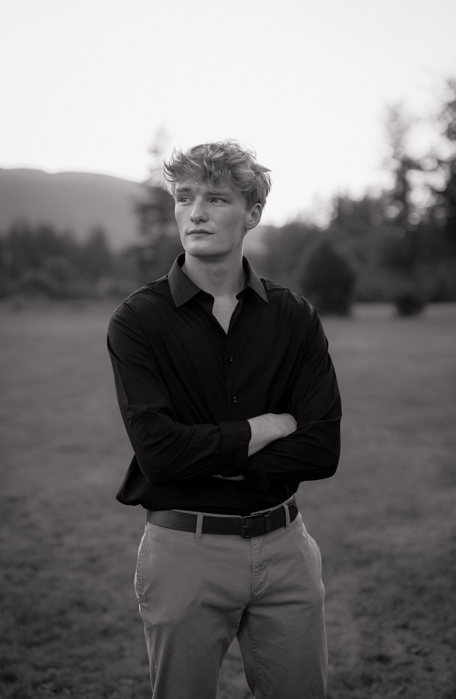 Young man with crossed arms in black shirt and khaki pants stands in a field, black and white portrait photography.