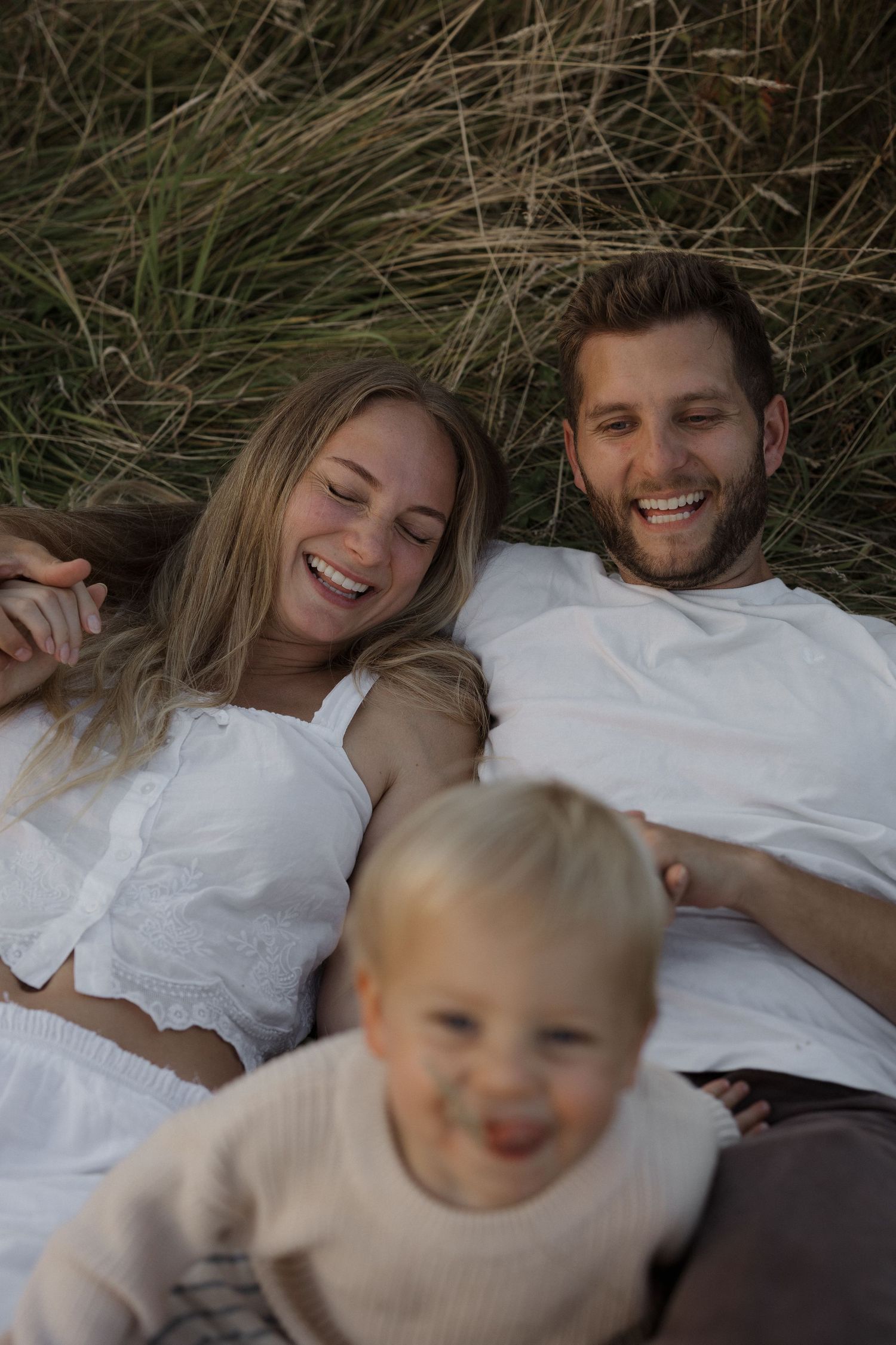 Happy family laughing outdoors in tall grass, parents lying down with young toddler child in foreground.