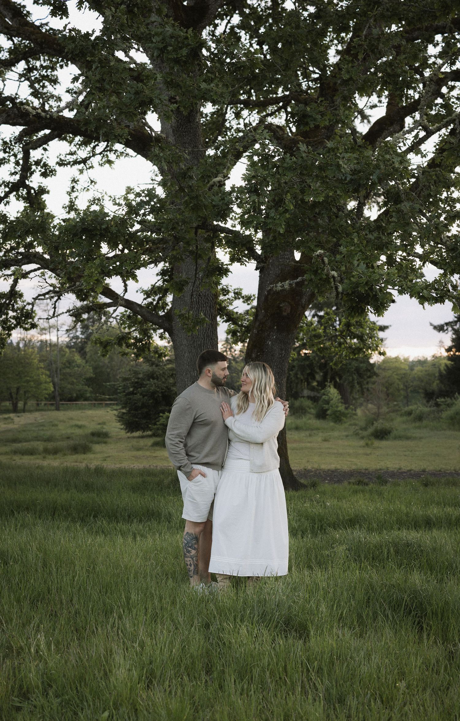 Couple embracing in a lush green field beneath a large oak tree during golden hour.