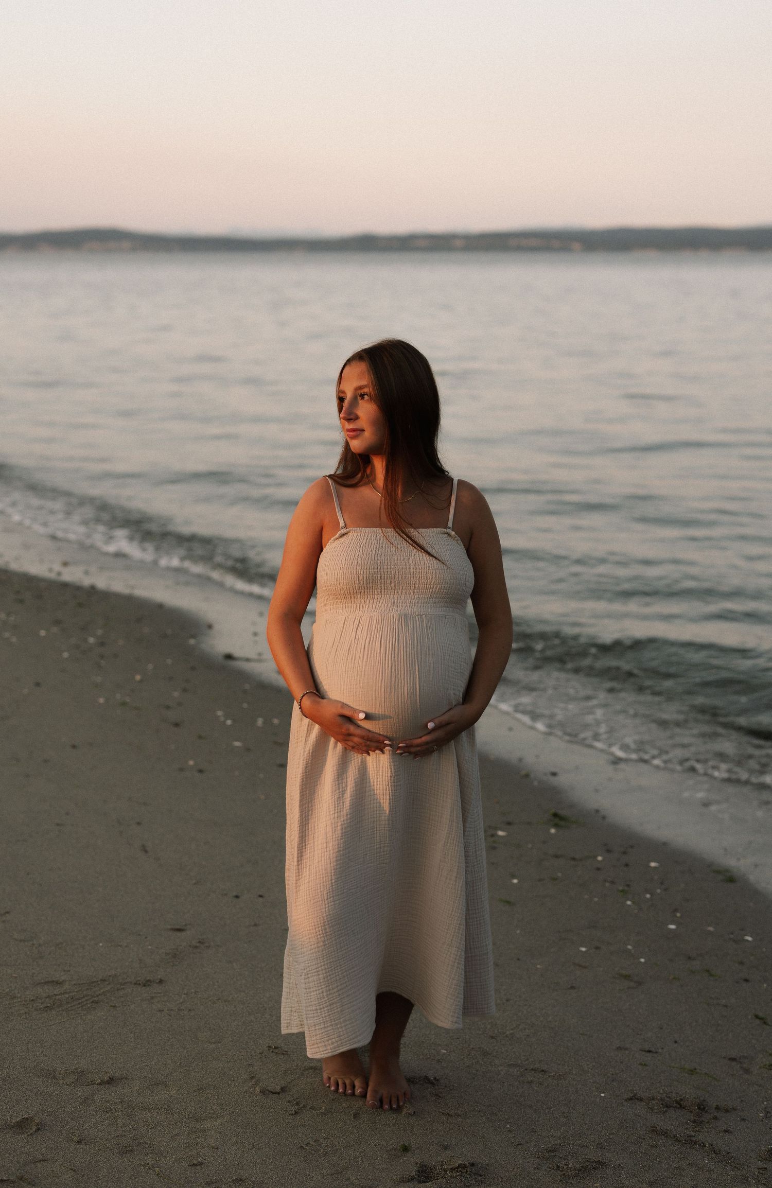 Pregnant woman in white dress cradling her baby bump on a sandy beach at sunset.