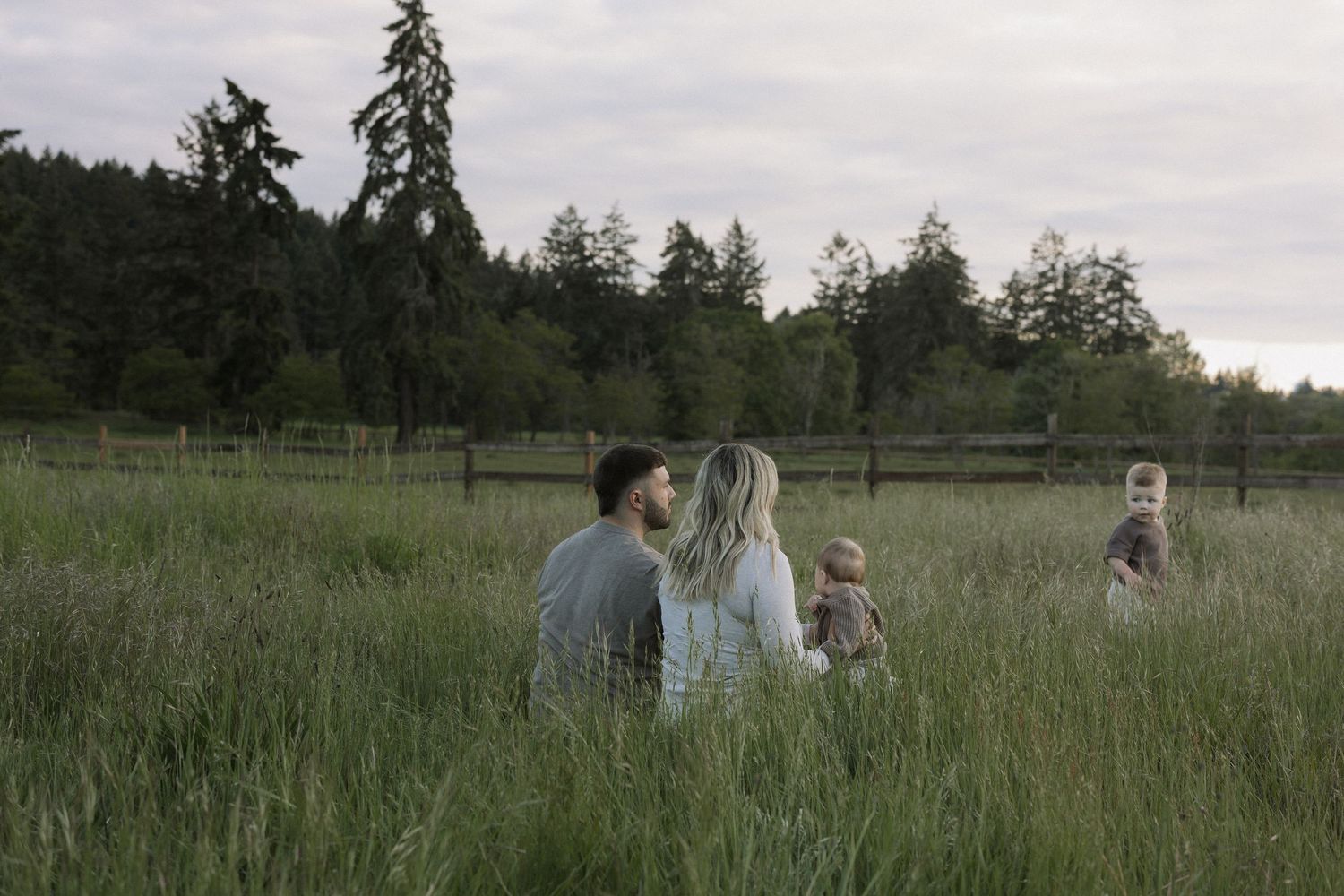A family sits together in a lush green field surrounded by tall trees and a wooden fence at dusk.