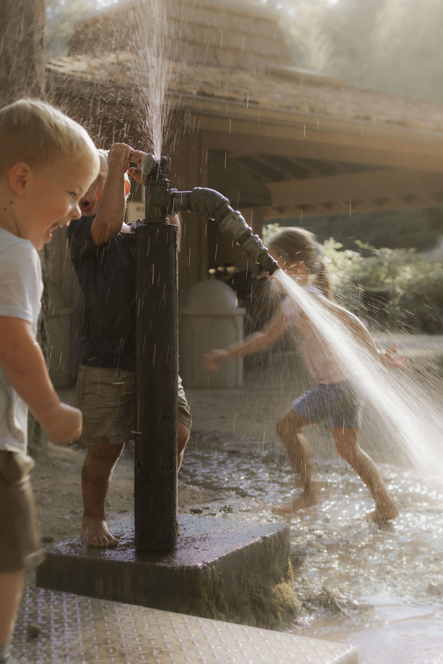 Children playing at an outdoor water pump fountain on a sunny day, splashing and laughing in the water.