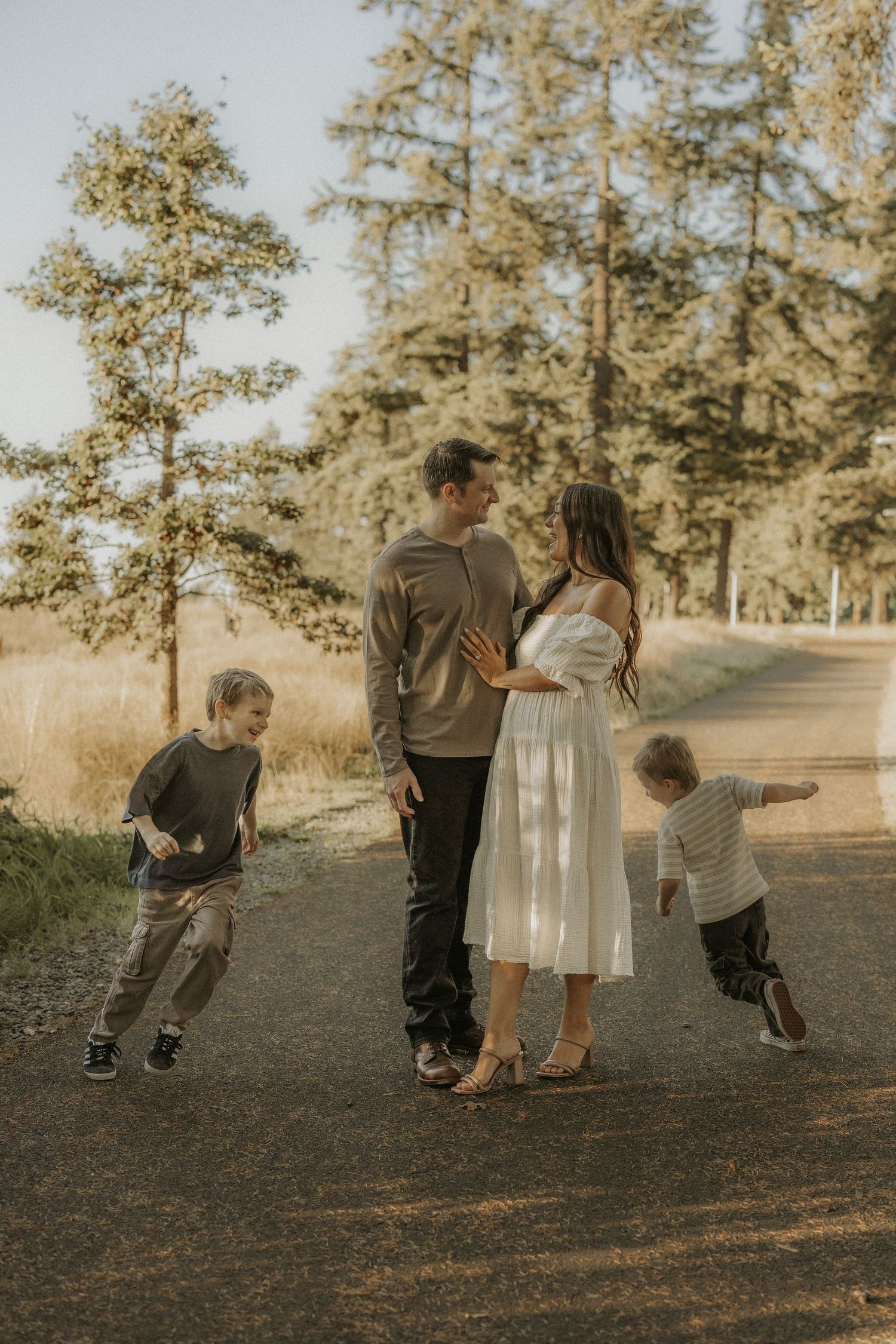 Family of four on a scenic path surrounded by tall trees, two playful boys flanking their parents in a warm outdoor setting.