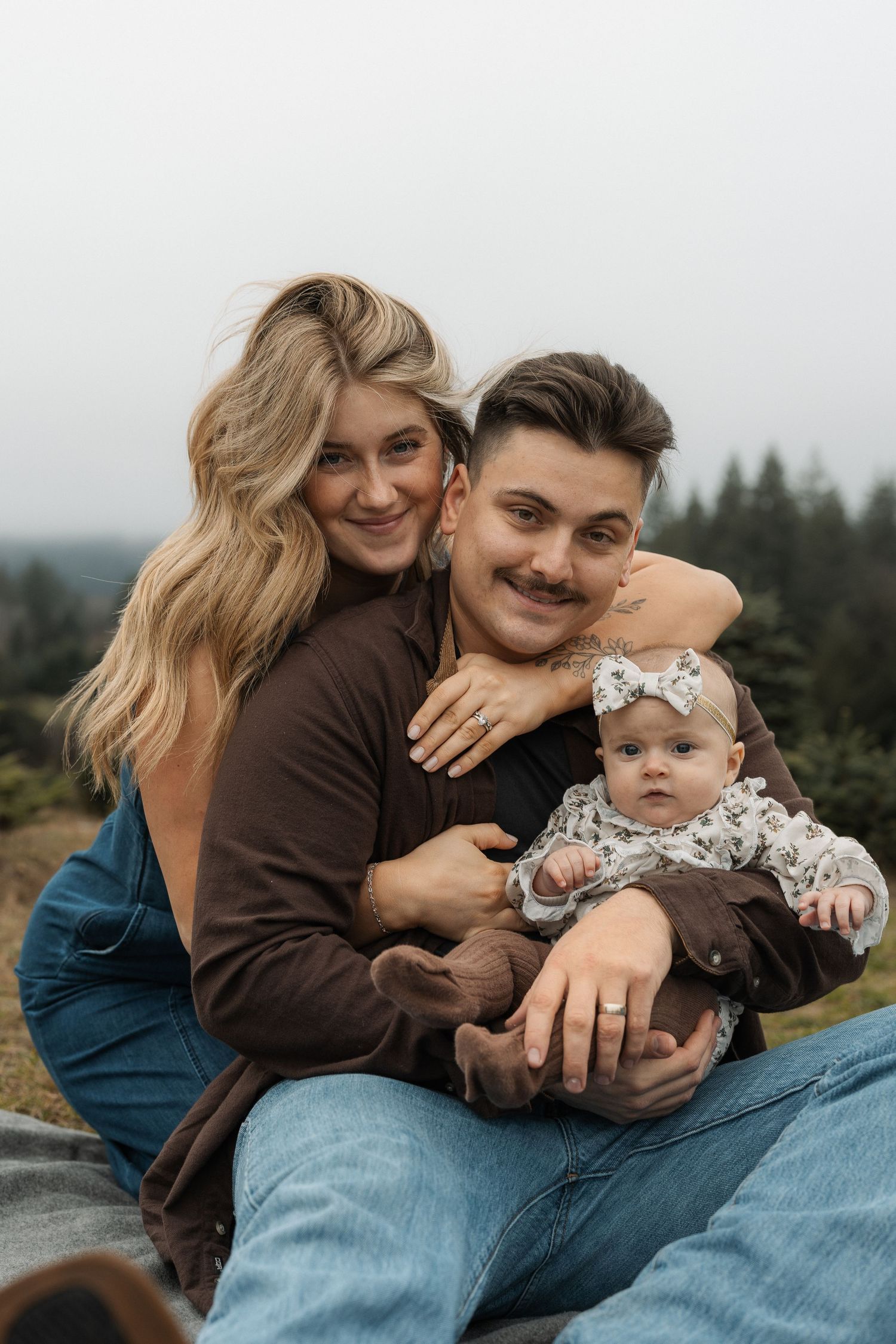 A young couple sitting outdoors holding their baby girl, smiling warmly with a forested hillside in the background.