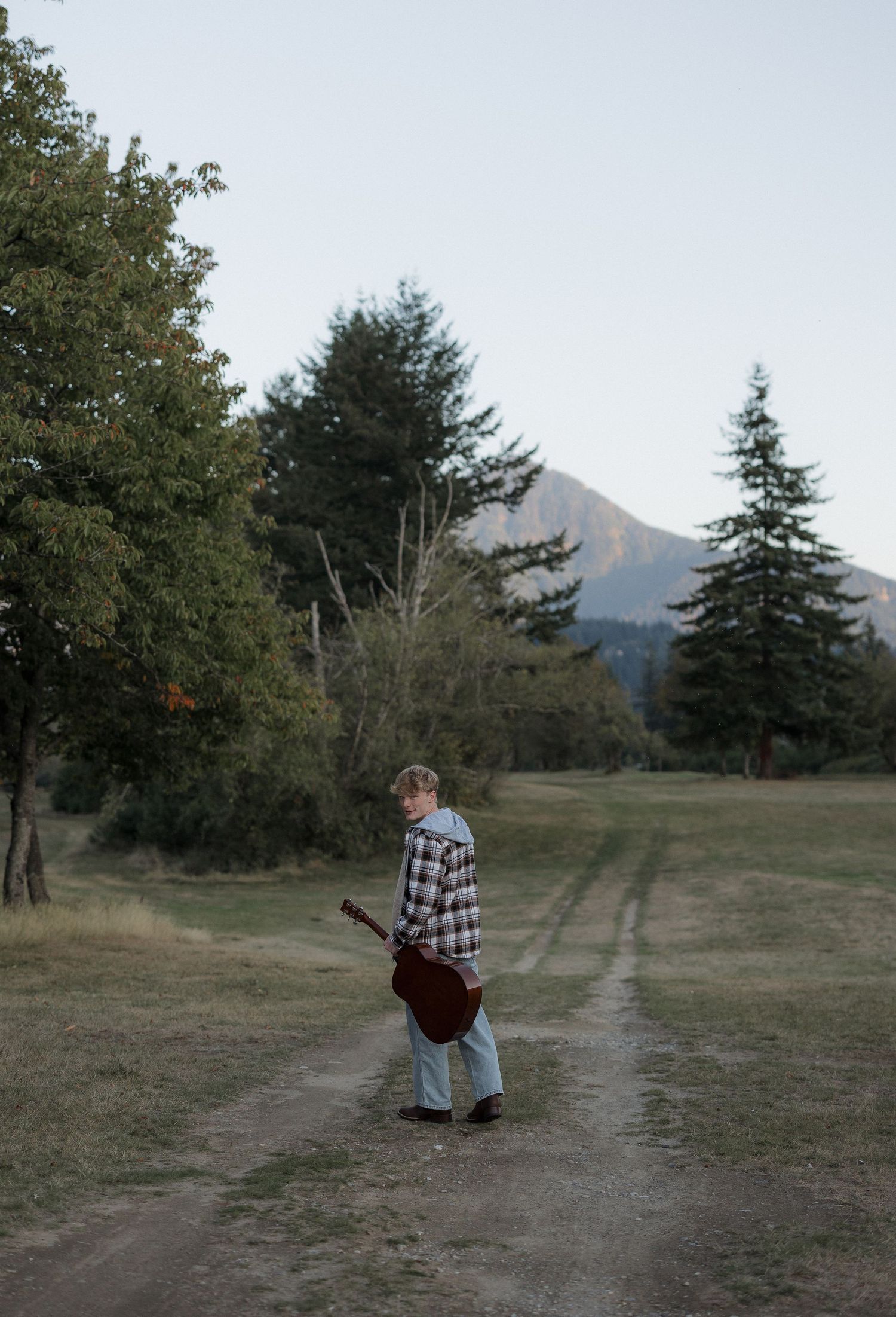 Person holding a guitar walking on a dirt path surrounded by trees with a mountain in the background.