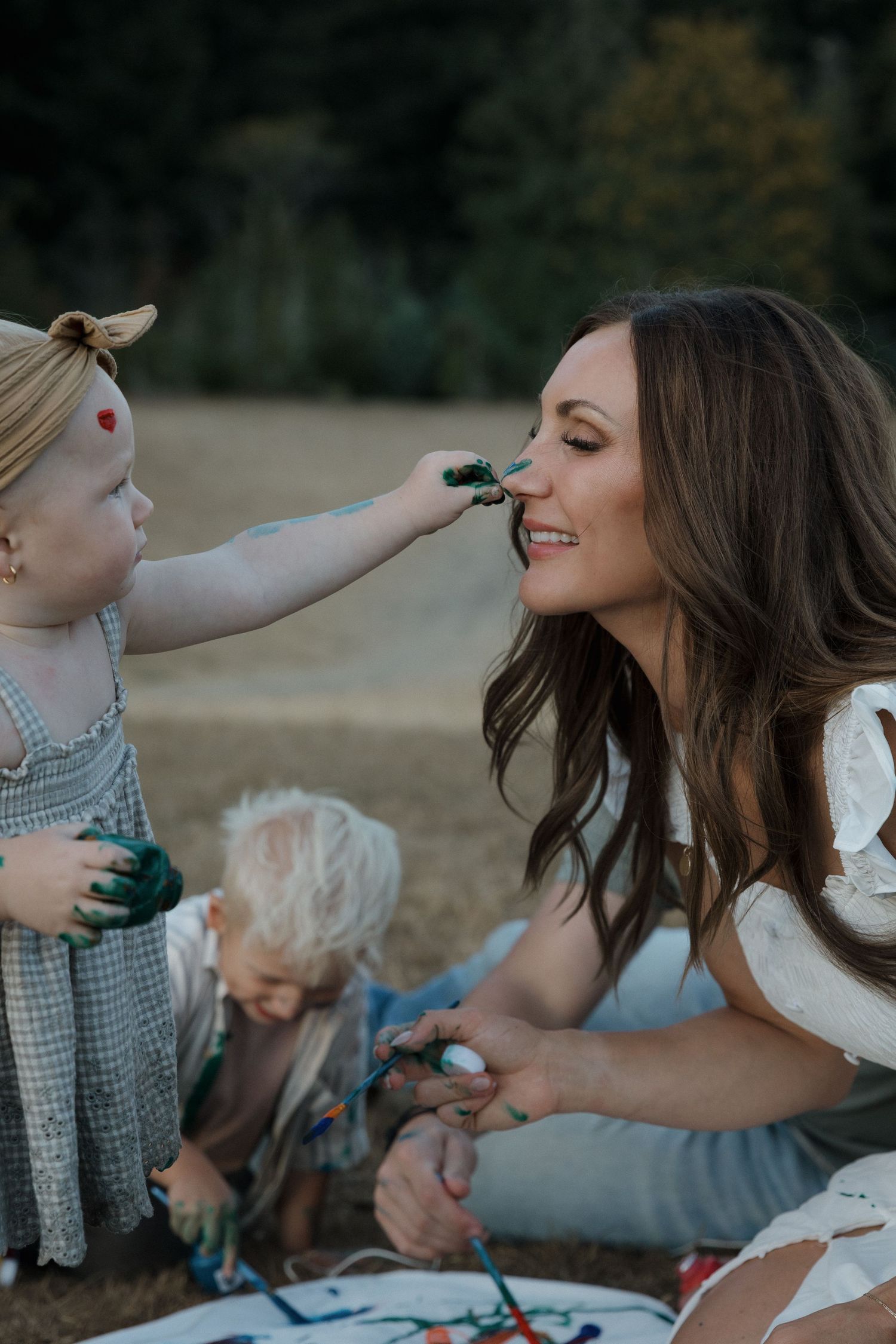 A toddler girl playfully paints her smiling mother's nose green during an outdoor family art session.