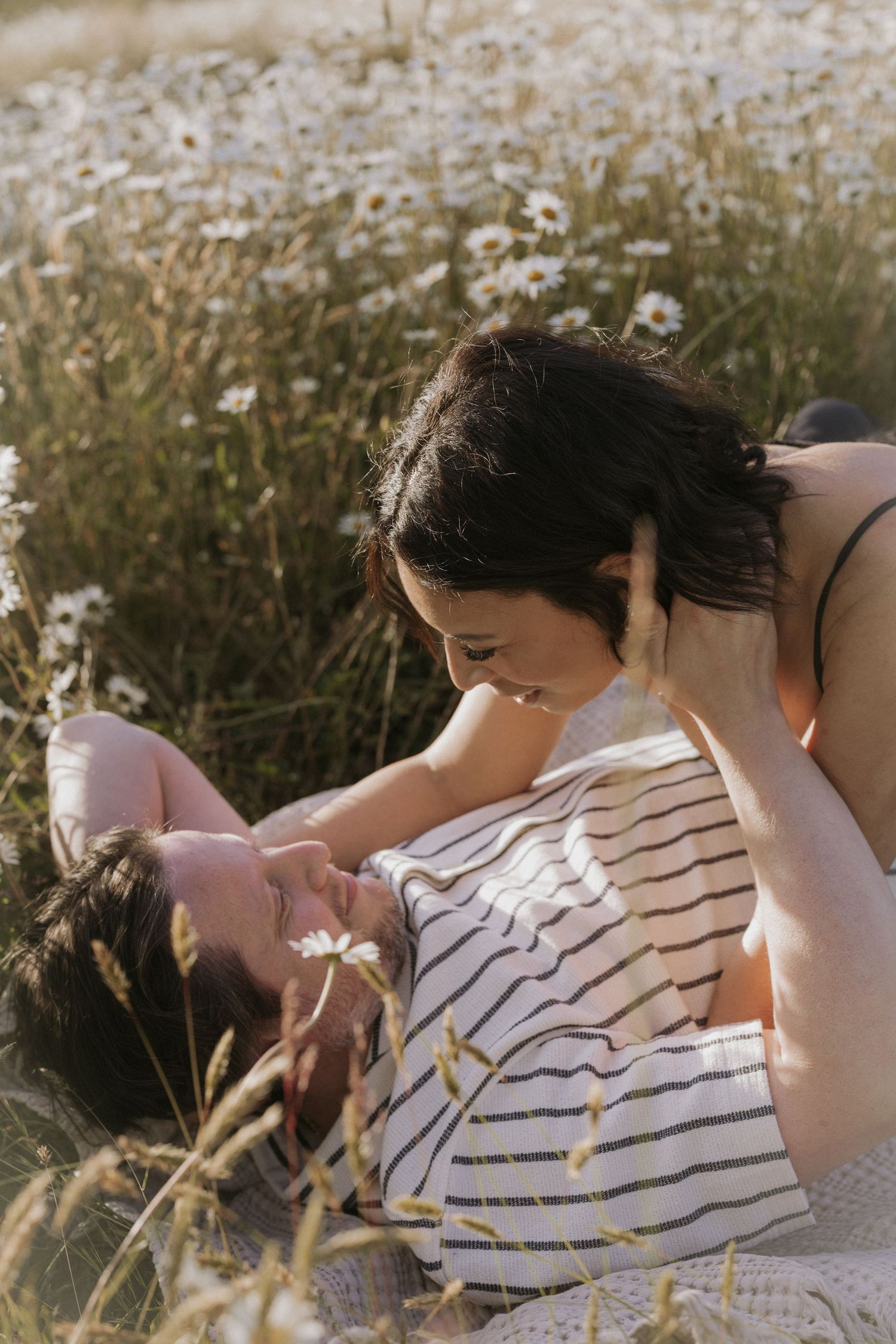 Two women share a romantic moment in a sunlit daisy field, one lying down as the other leans in close.