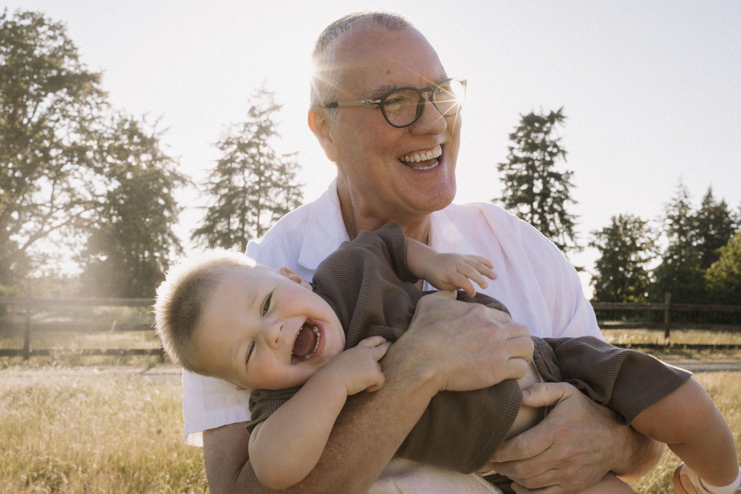 Laughing grandfather holding giggling toddler boy outdoors in sunny park with green trees in background.