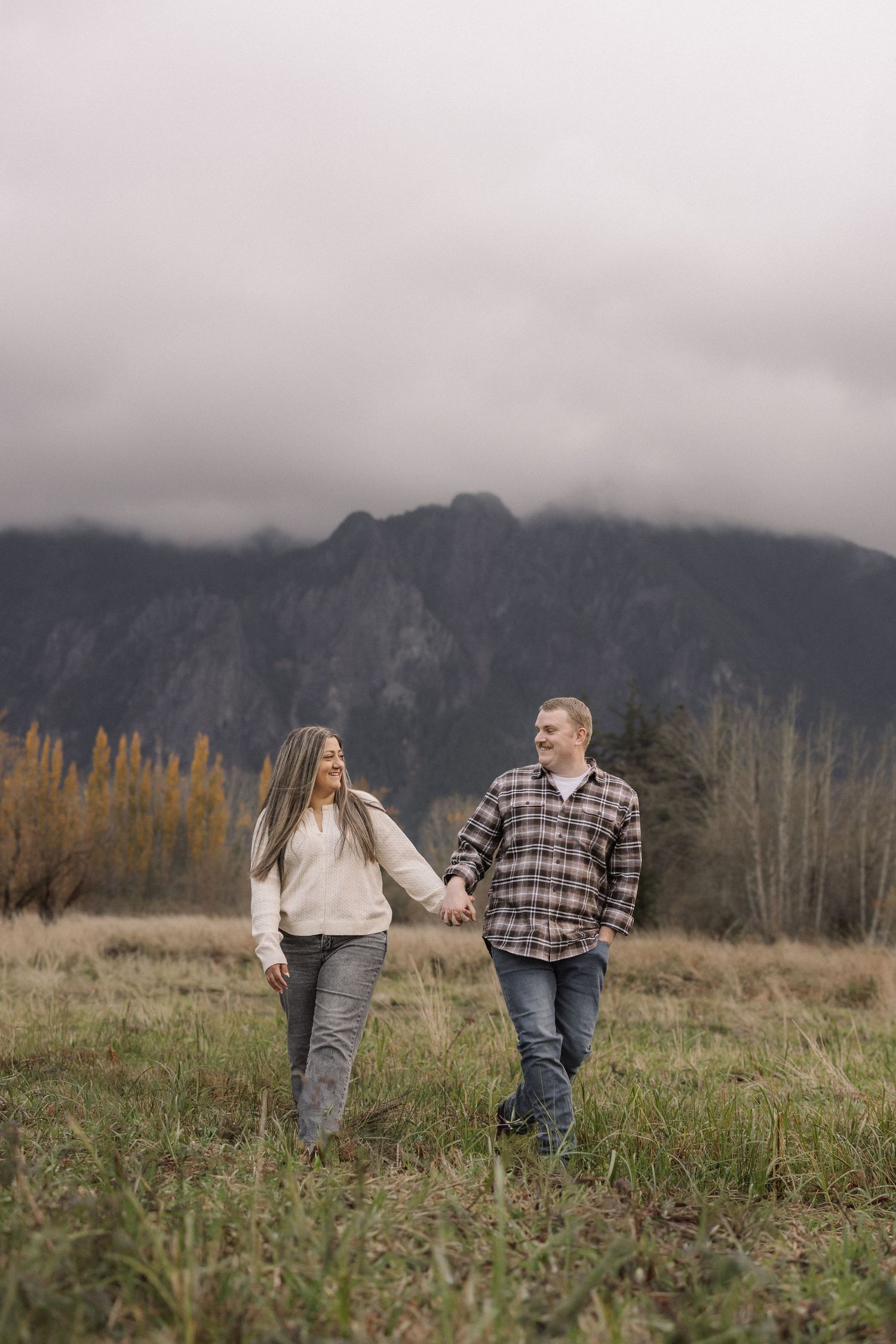 Couple holding hands walking through a grassy field with misty mountains in the background on a cloudy day.