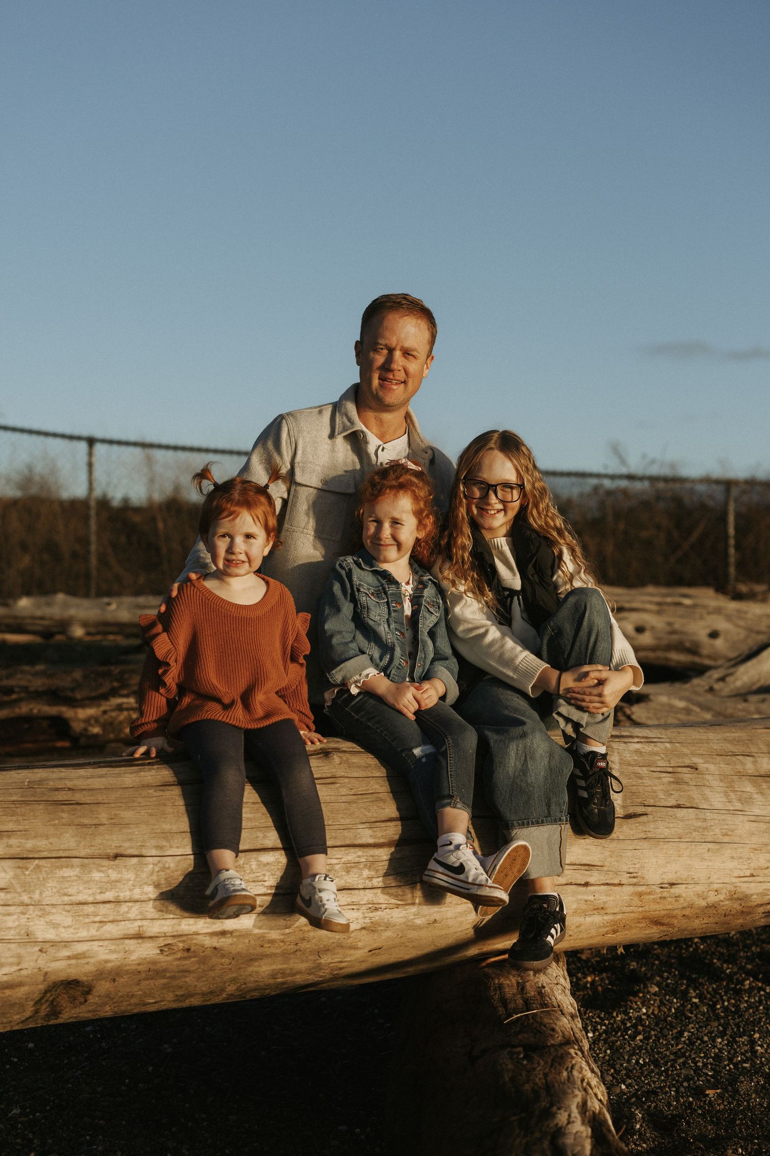 Family of four posing on a large log outdoors during golden hour, with two redhead children and parents smiling.
