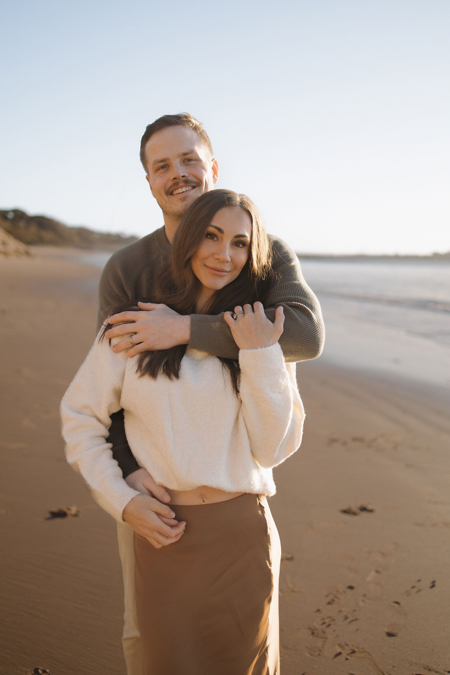 A couple embracing on a sandy beach at golden hour, smiling warmly in cozy sweaters with ocean waves behind them.