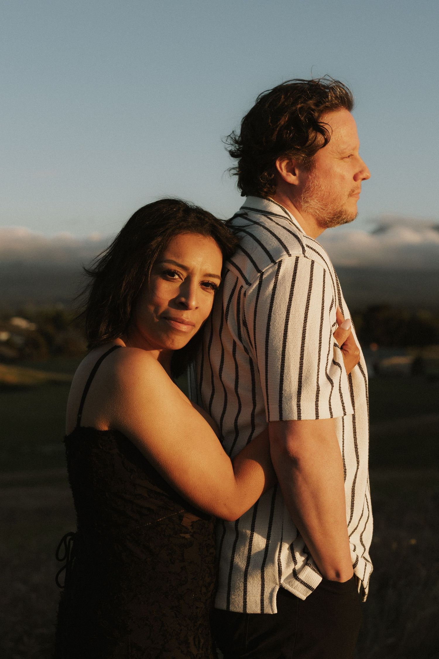 A woman in a black dress embraces a man in a striped shirt from behind at golden hour outdoors.