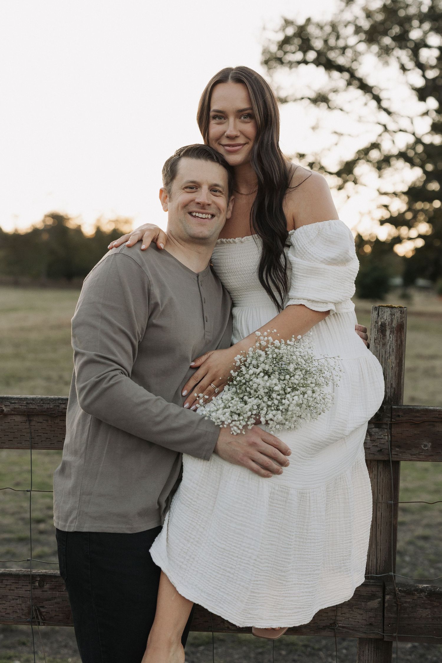 Happy couple posing outdoors at sunset, woman in white dress holding baby's breath bouquet, man in olive shirt embracing her.