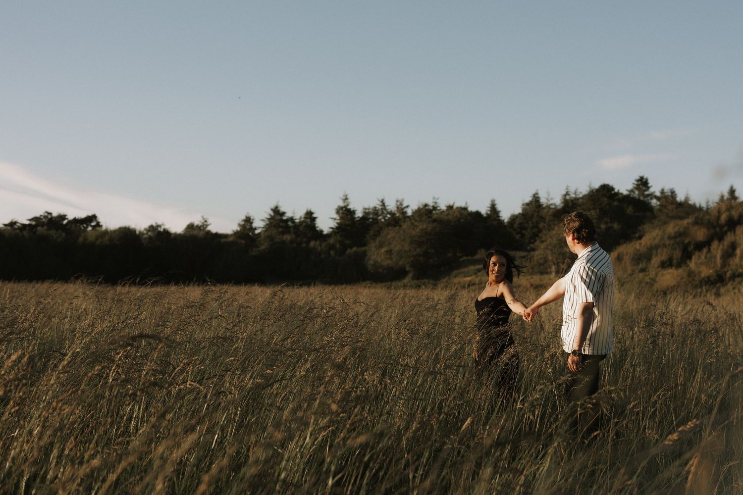 A couple holds hands walking through a golden grass field at dusk with trees in the background.