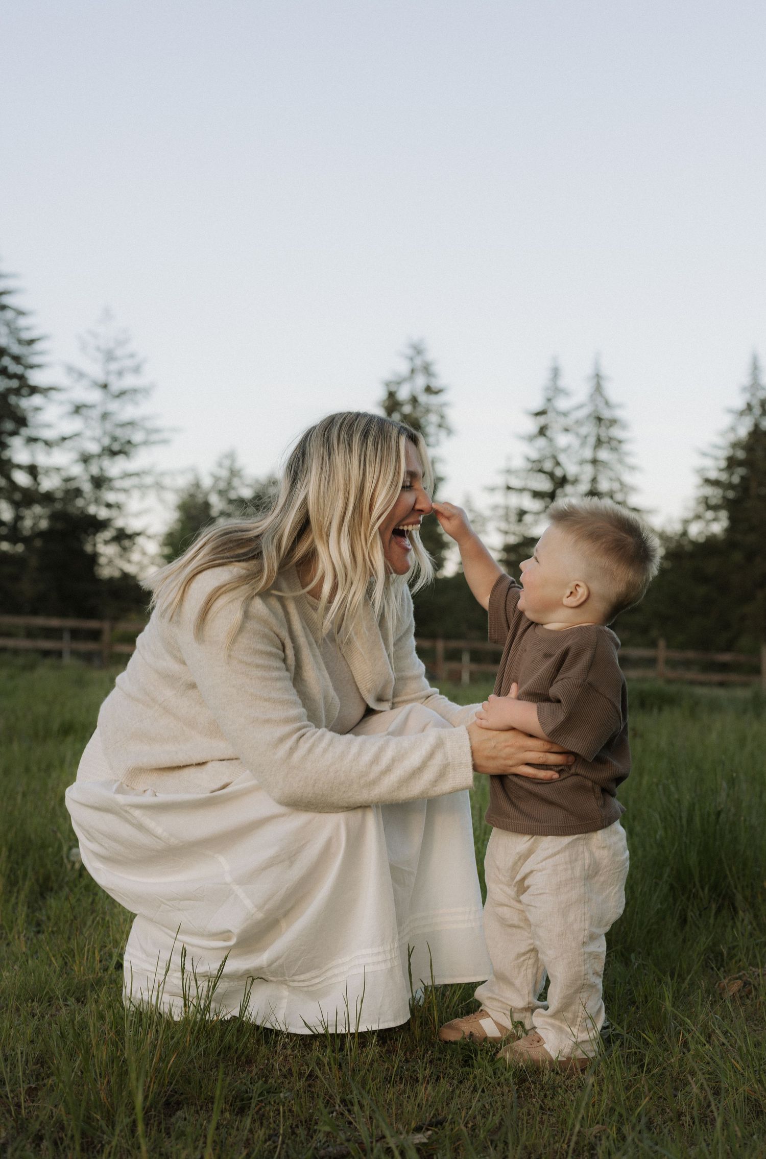 A blonde woman crouches in a green field, smiling as a toddler touches her nose, with tall trees in the background.