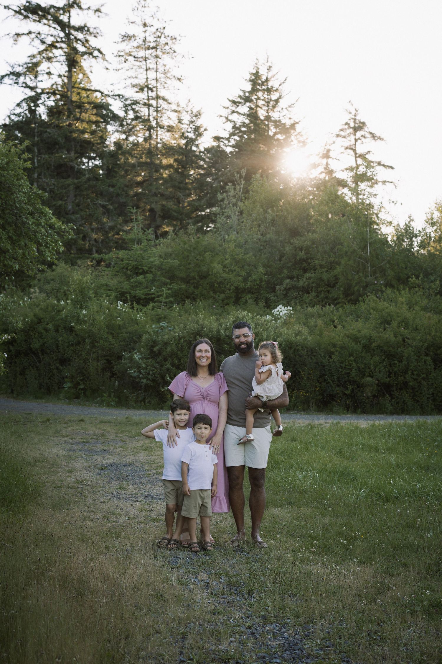 Family of five posing outdoors at golden hour with lush green trees in background.