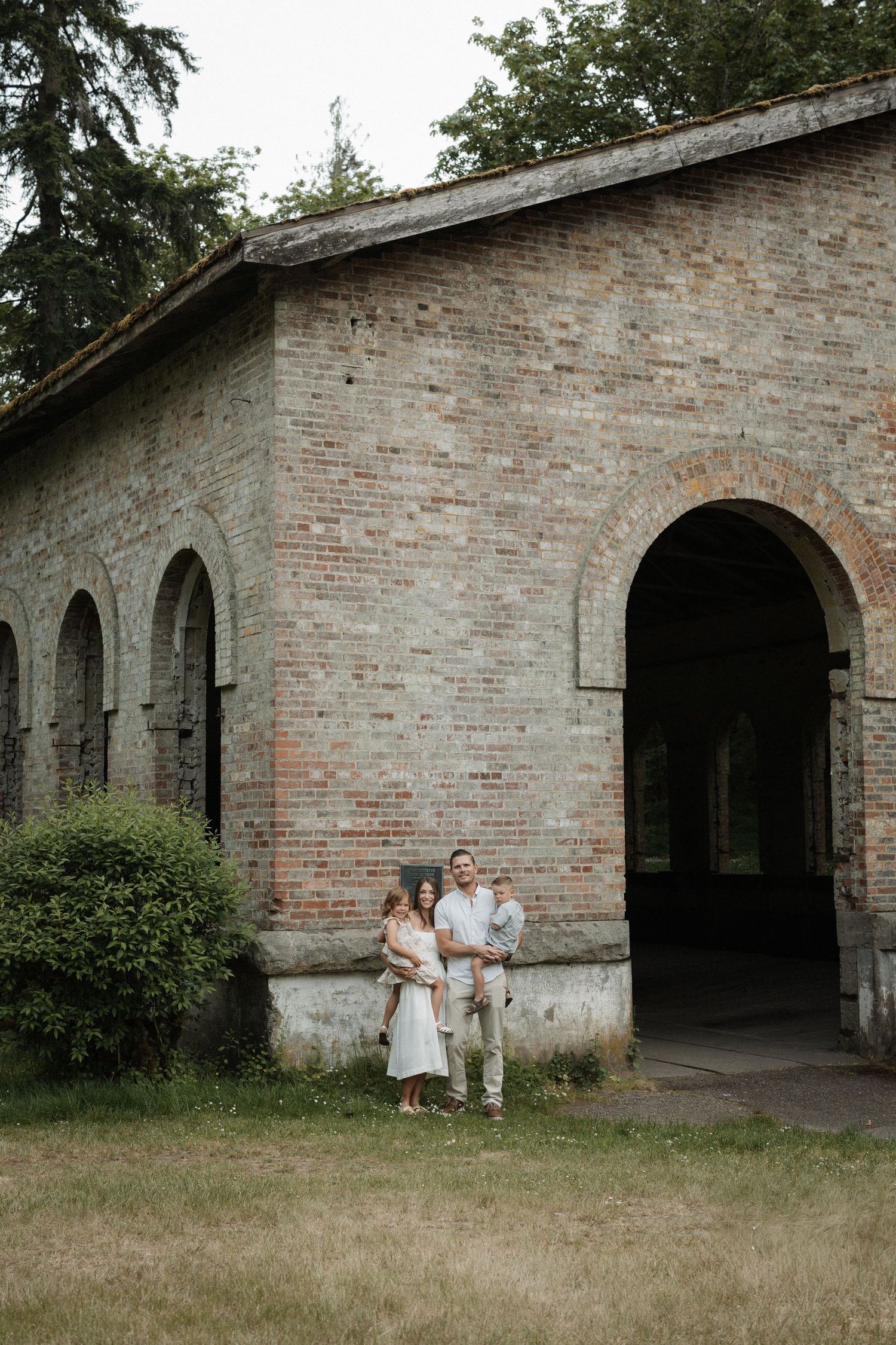 Family of three posing in front of a rustic brick building with arched windows and doorways surrounded by lush greenery.