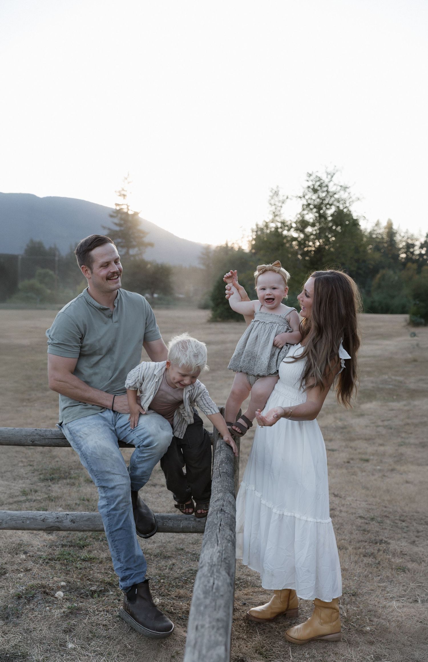 Family of four laughing outdoors at golden hour near a wooden fence with mountains and trees in the background.