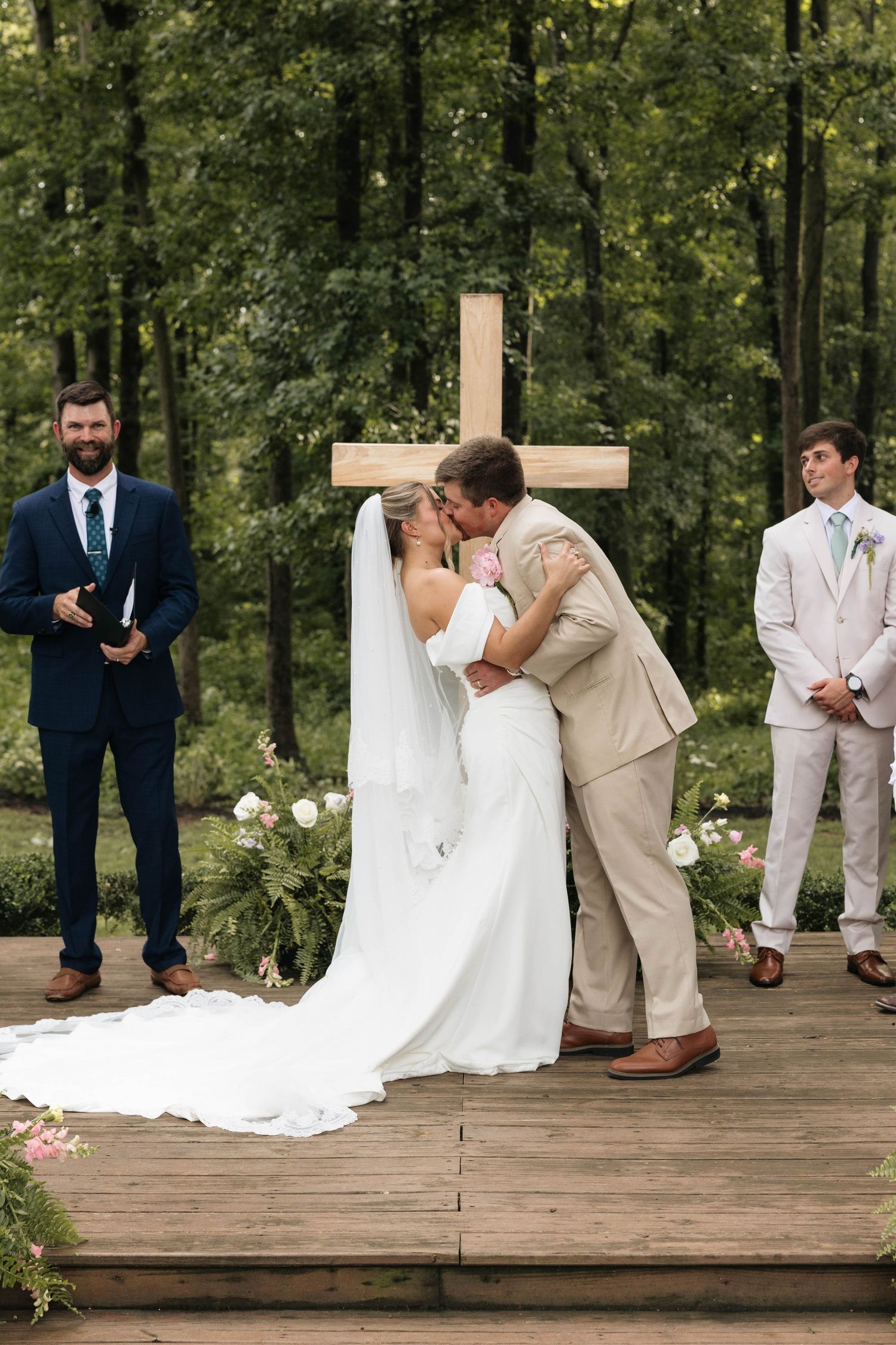 Bride and groom share first kiss at outdoor wedding ceremony with wooden cross backdrop surrounded by lush green trees.