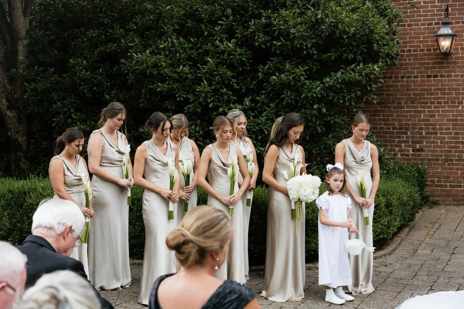 Bridesmaids in champagne satin gowns holding calla lilies stand with flower girl before ceremony.