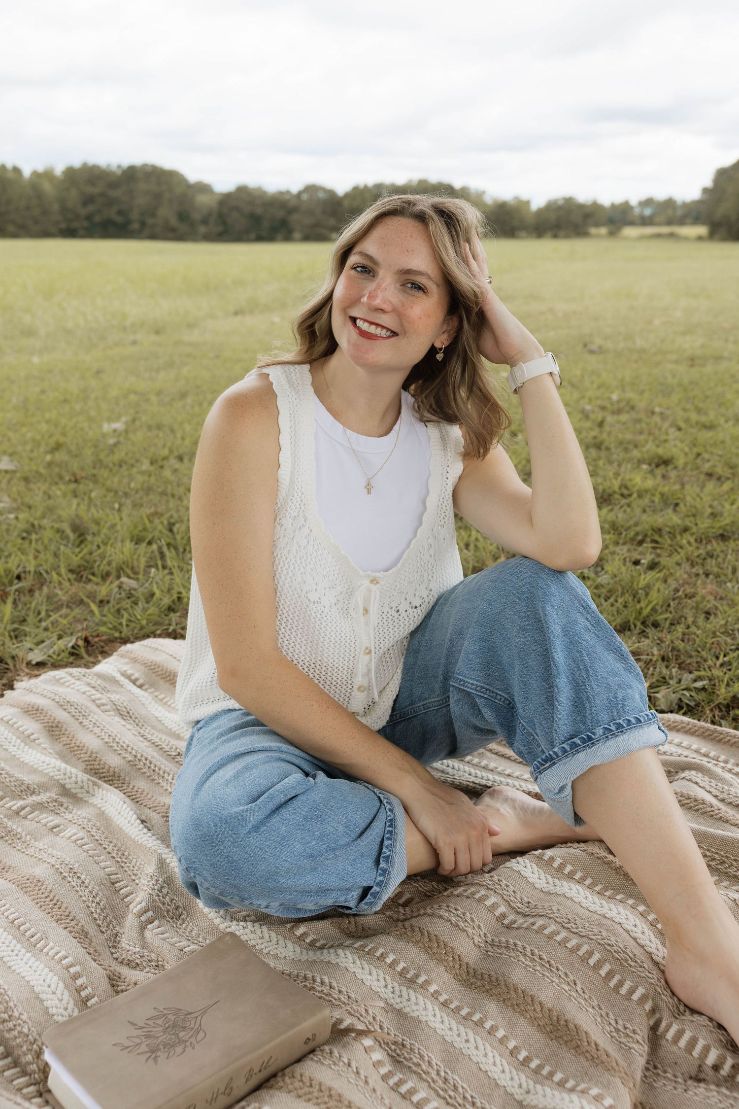 Young woman in white top and jeans sitting on blanket in green field, smiling and posing for portrait photo.