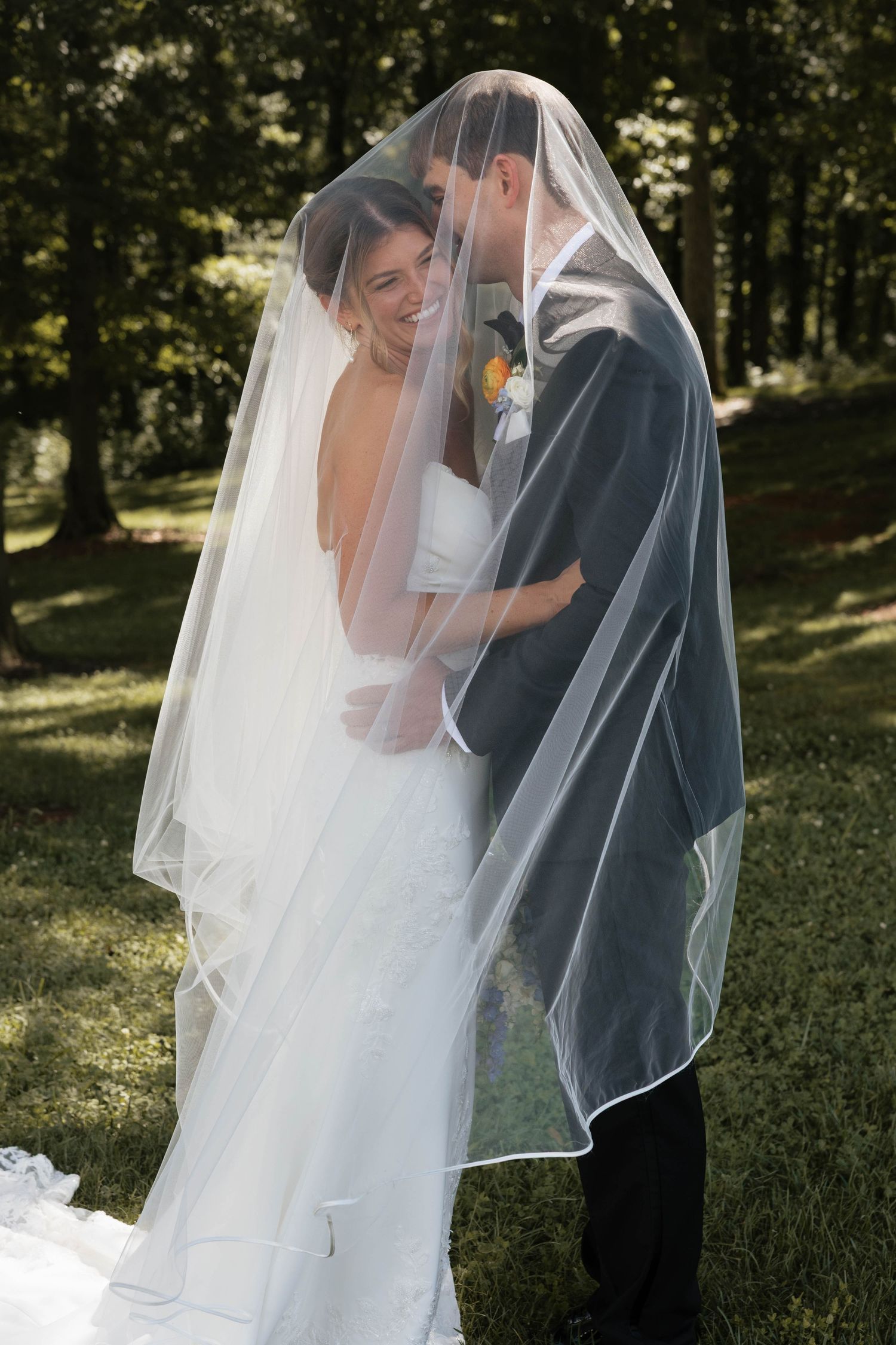 Bride and groom sharing romantic moment under flowing white veil in lush green outdoor setting.
