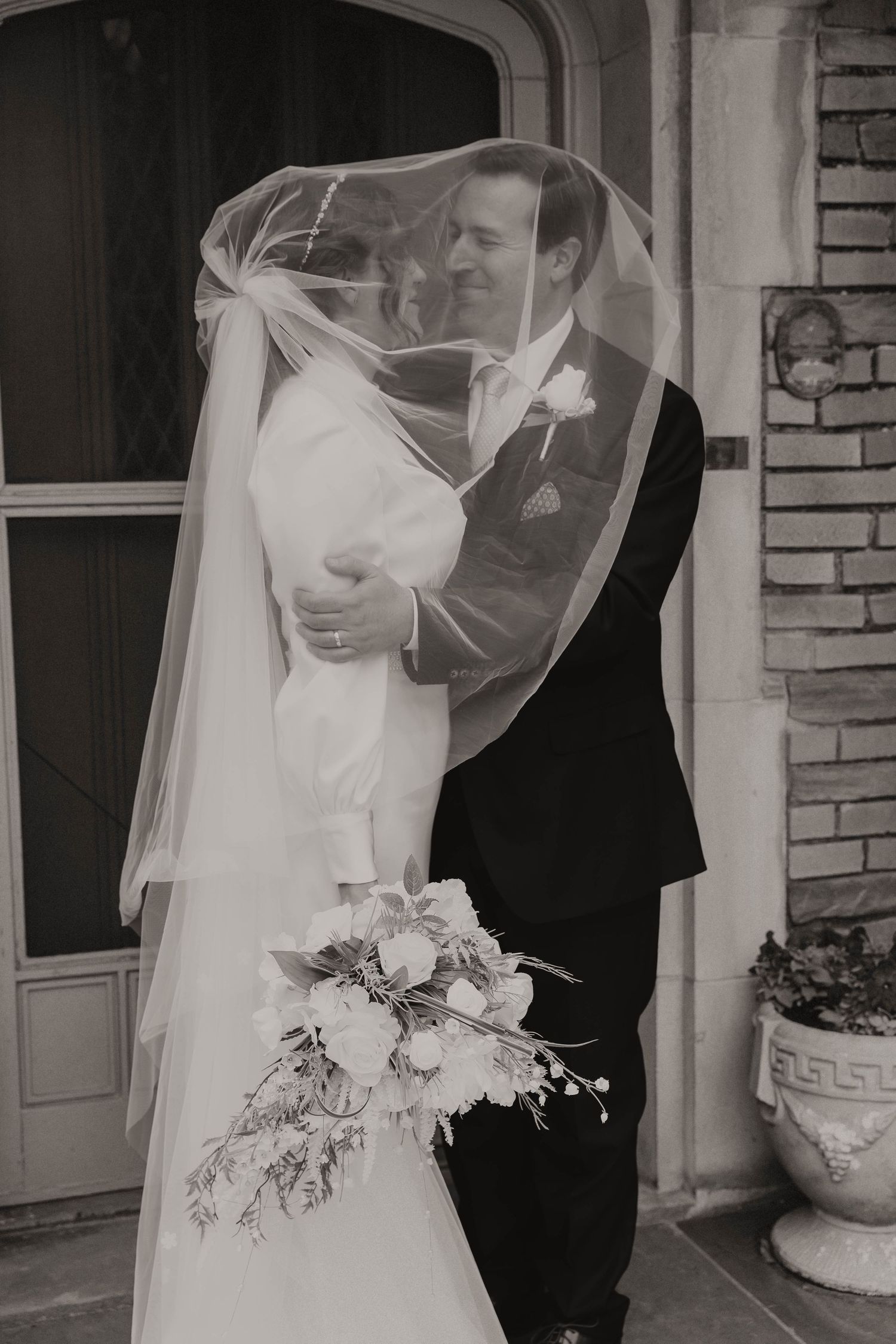 Bride and groom share romantic moment under veil holding white bouquet in black and white photo.