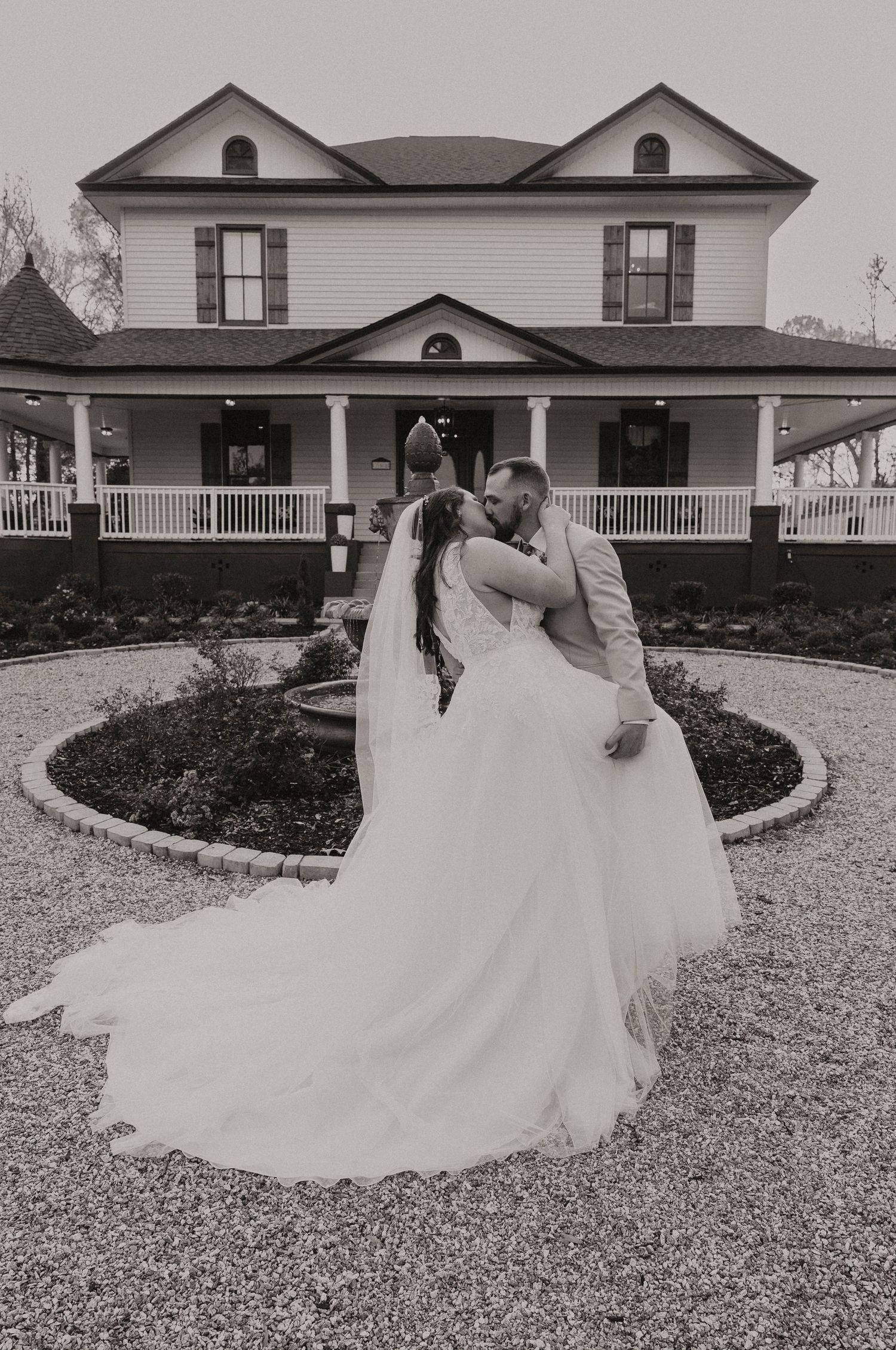 Black and white photo of bride and groom kissing in front of a classic white Southern manor house.