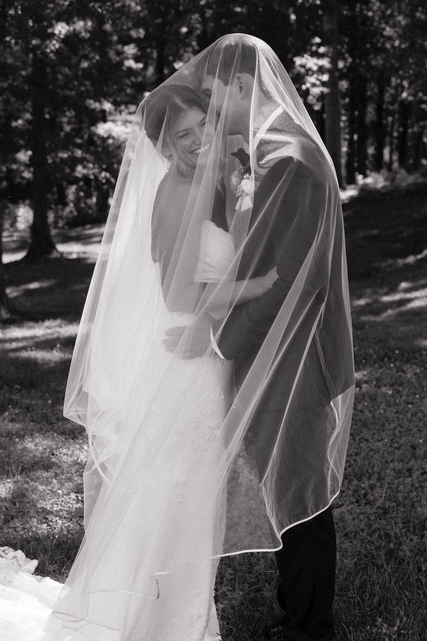 Black and white photo of bride and groom laughing together hidden beneath flowing wedding veil outdoors.