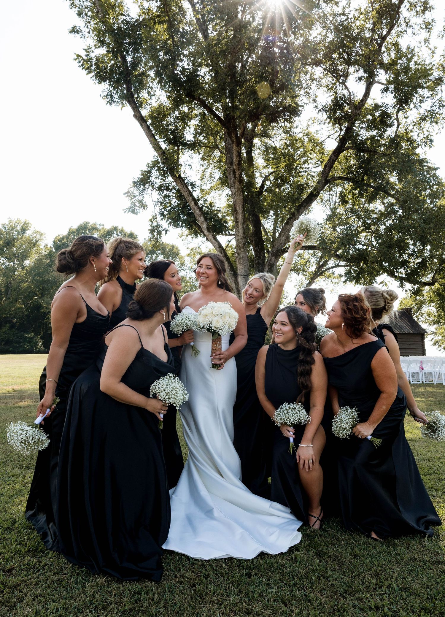 Bride in white gown with bridesmaids in black dresses holding baby's breath bouquets outdoors under a sunlit tree.