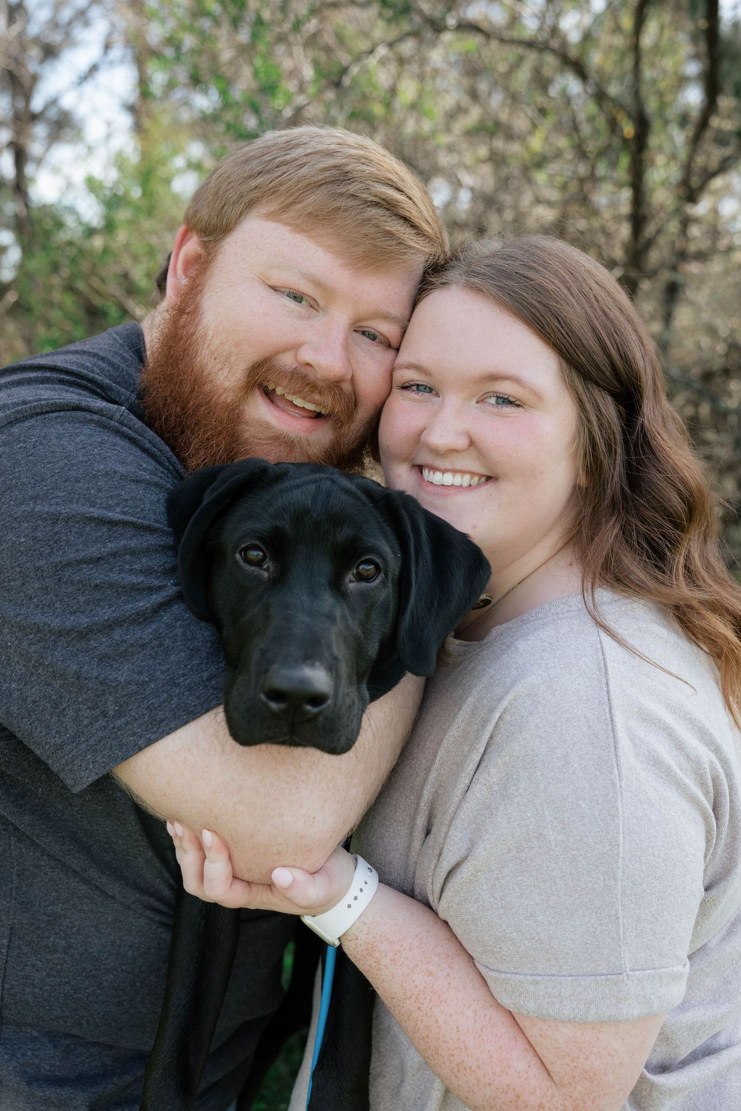 Smiling couple with red beard and brown hair hugging a black Labrador dog outdoors in a wooded setting.