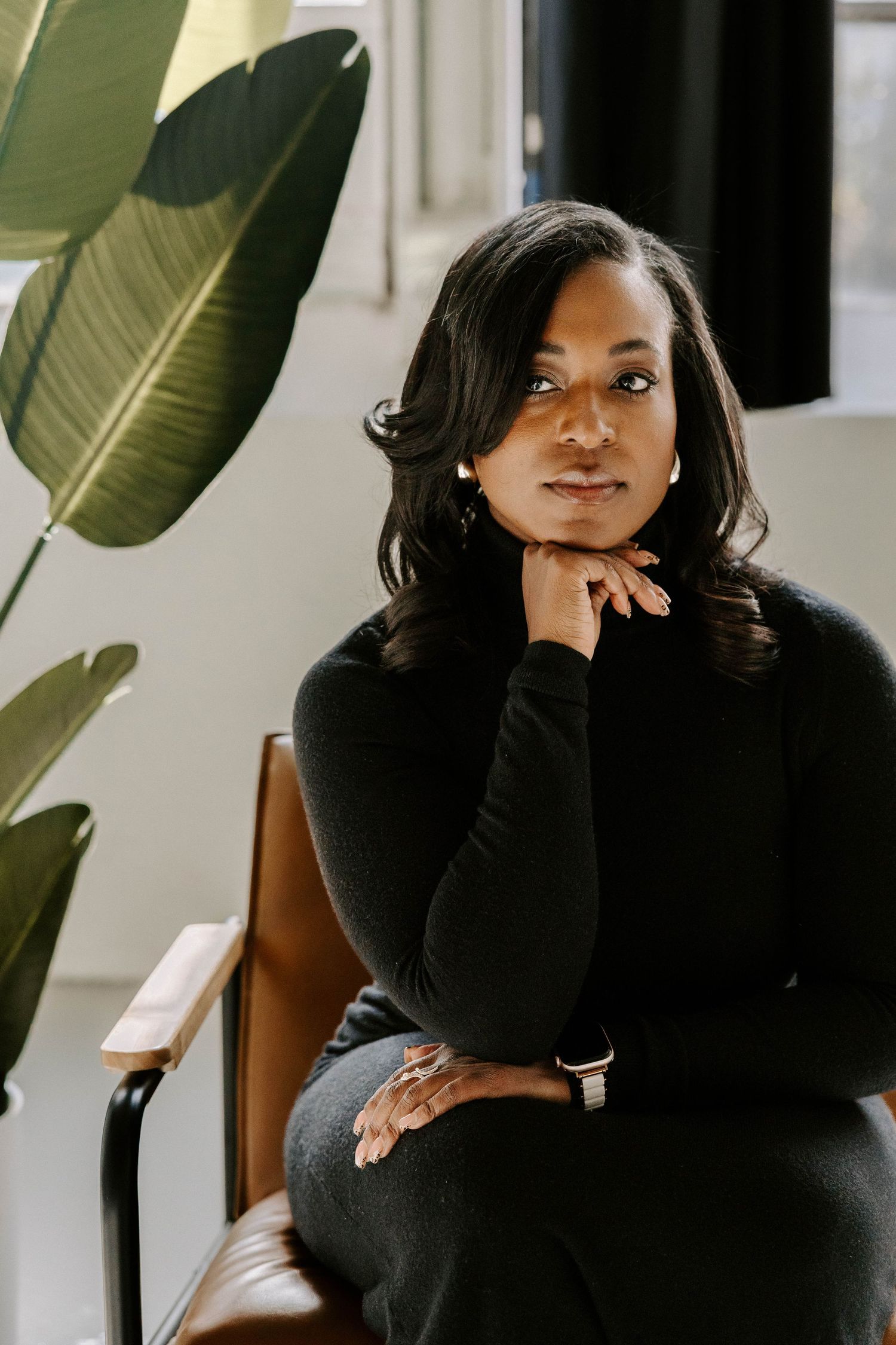 A woman in a black turtleneck sits thoughtfully in a chair beside a tropical plant near a bright window.