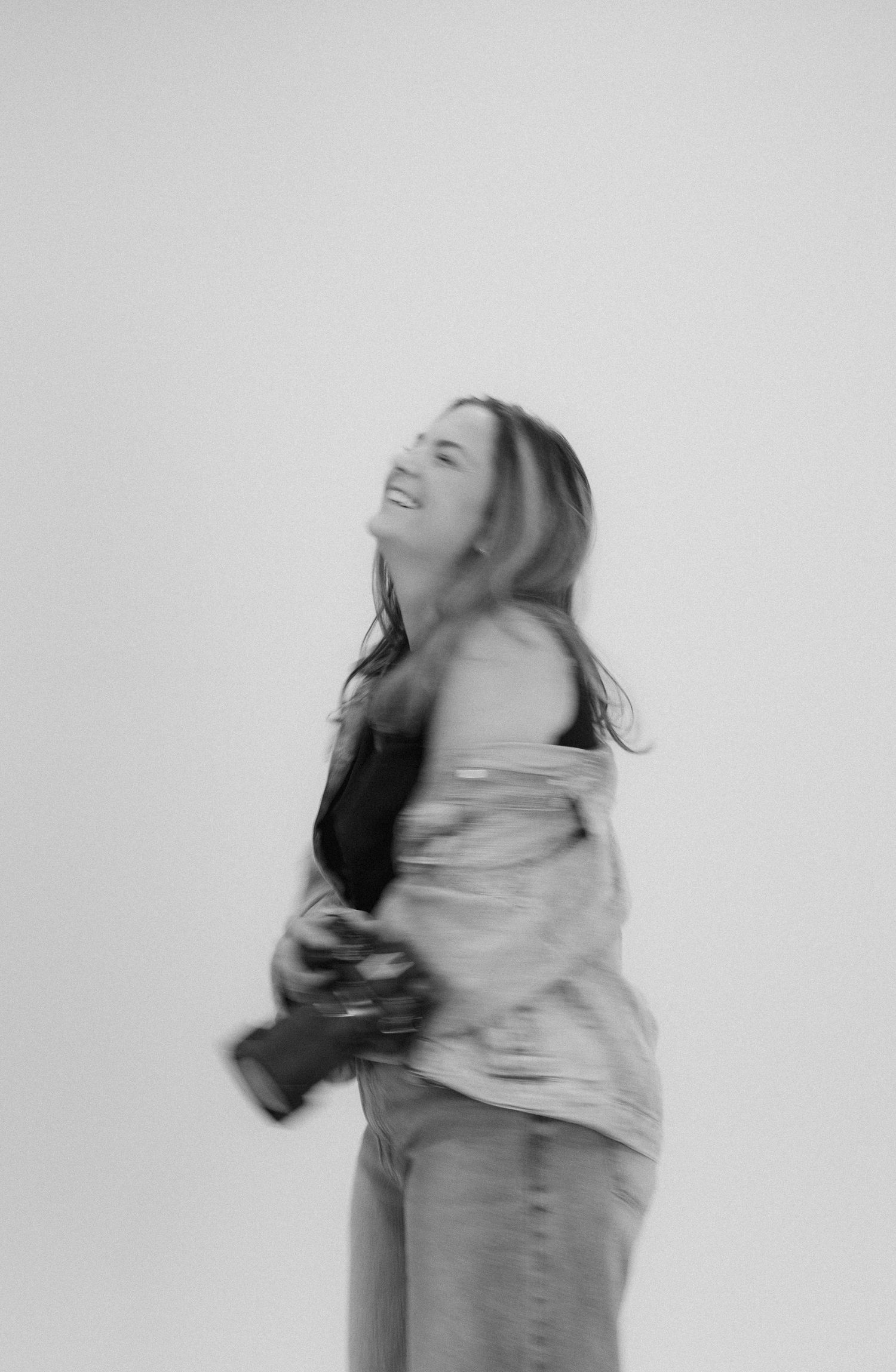 Black and white photo of a laughing woman holding a camera, captured with motion blur against a light background.