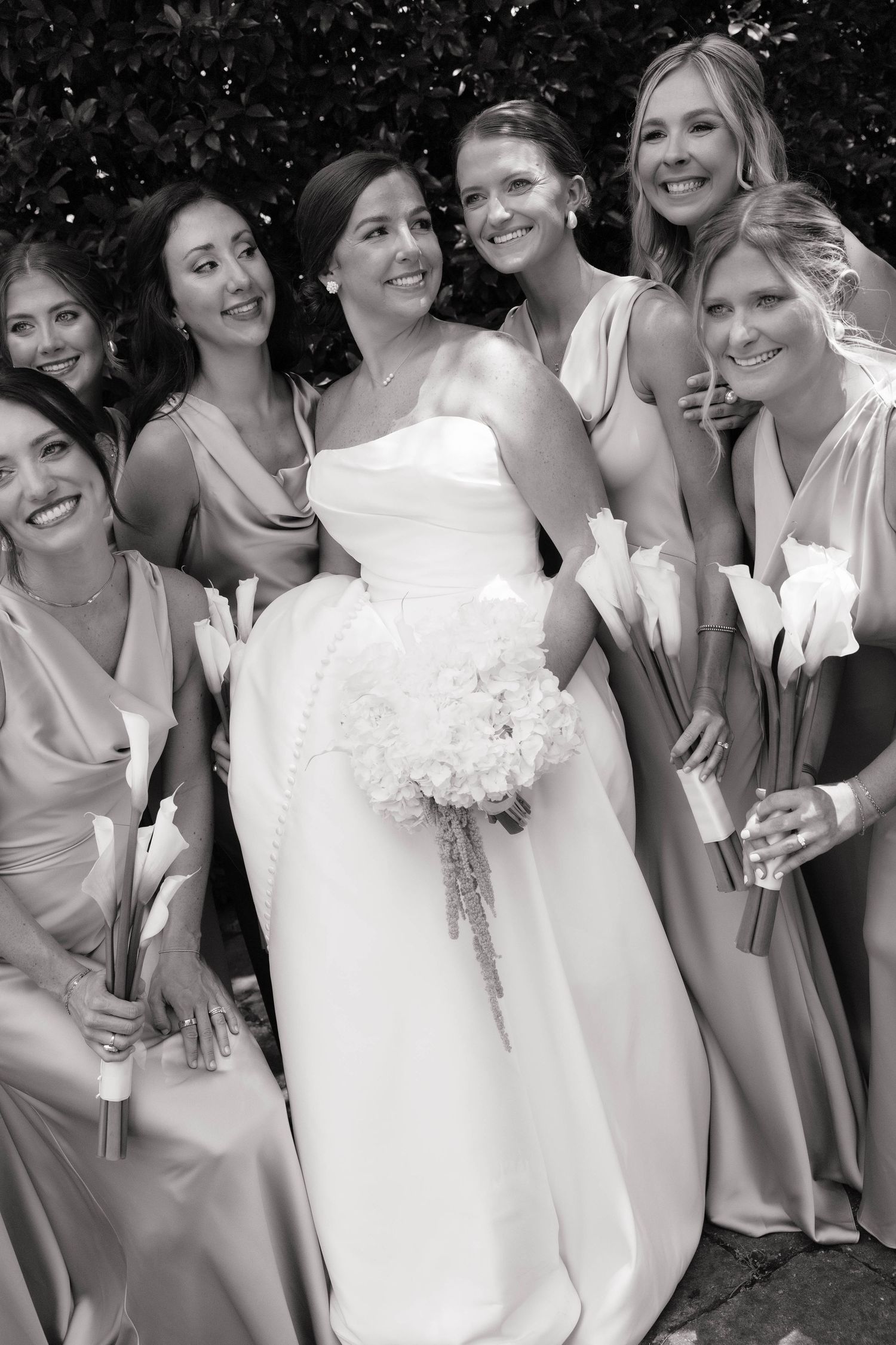 Black and white photo of bride with bouquet surrounded by smiling bridesmaids in elegant satin gowns.