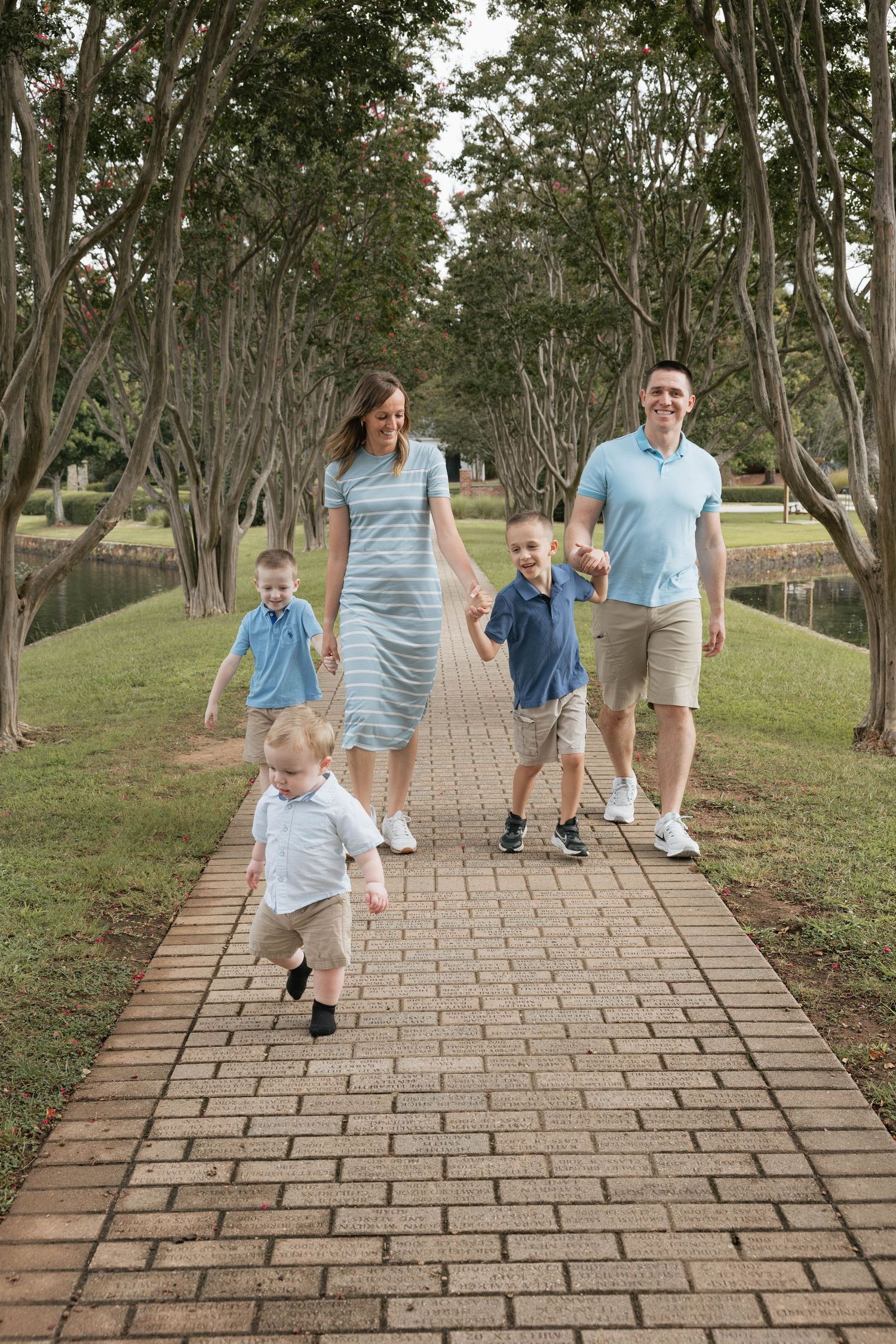 A happy family of five walks along a brick path lined with trees in a park, wearing coordinating blue outfits.