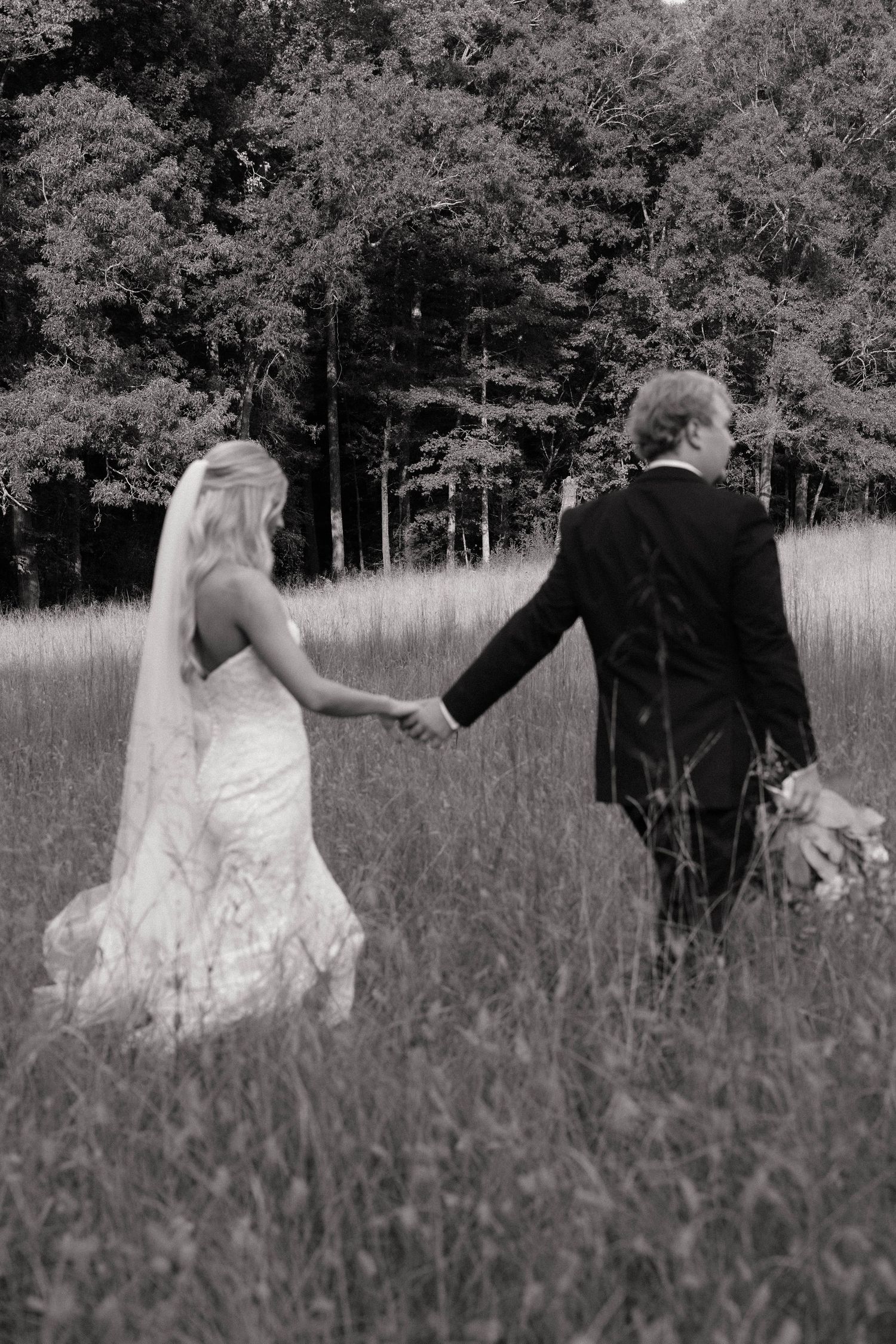 Black and white photo of bride and groom holding hands walking through a meadow toward a forest on their wedding day.