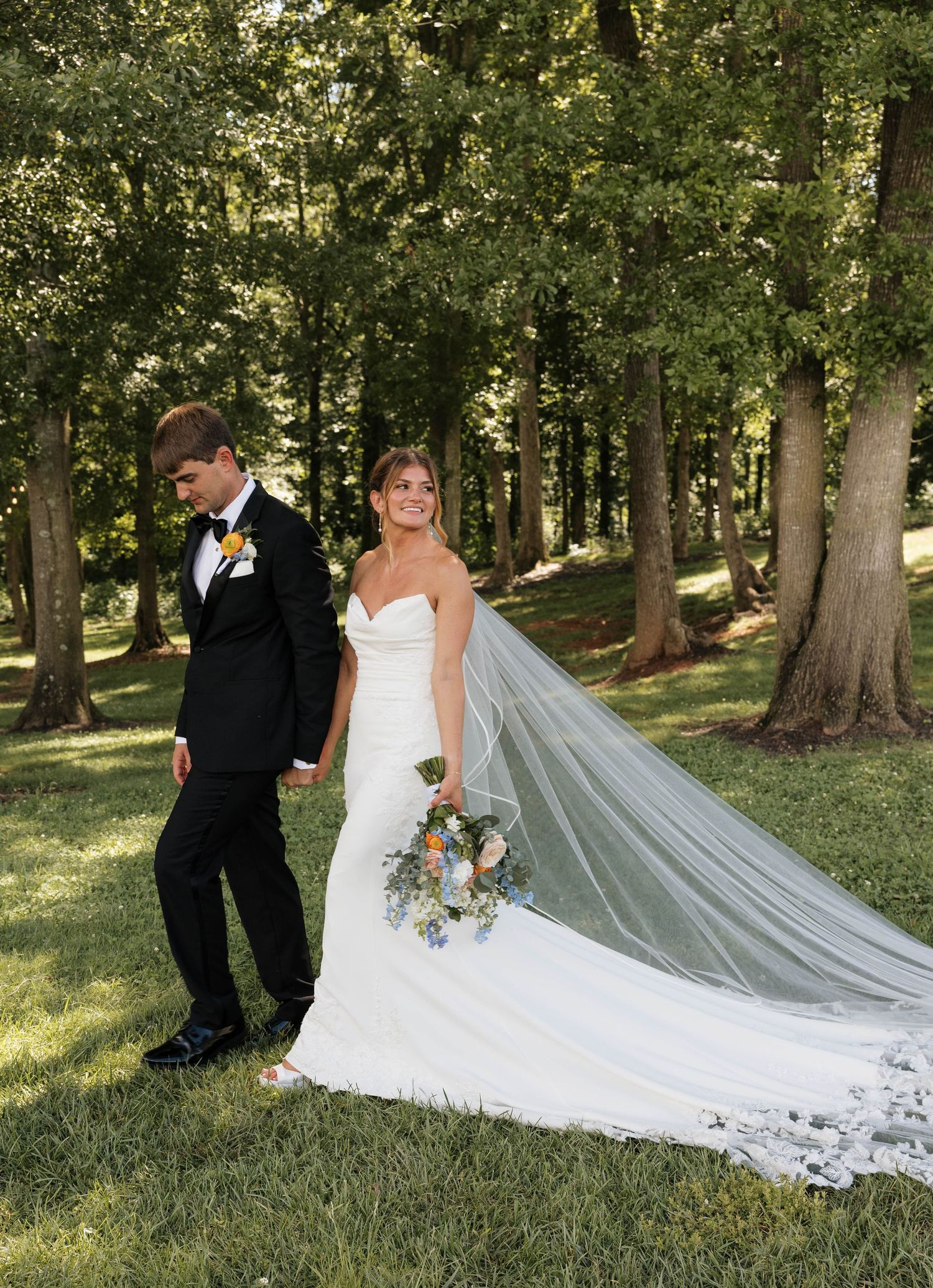 Bride in strapless gown holds bouquet while groom in black tuxedo poses among tall trees.