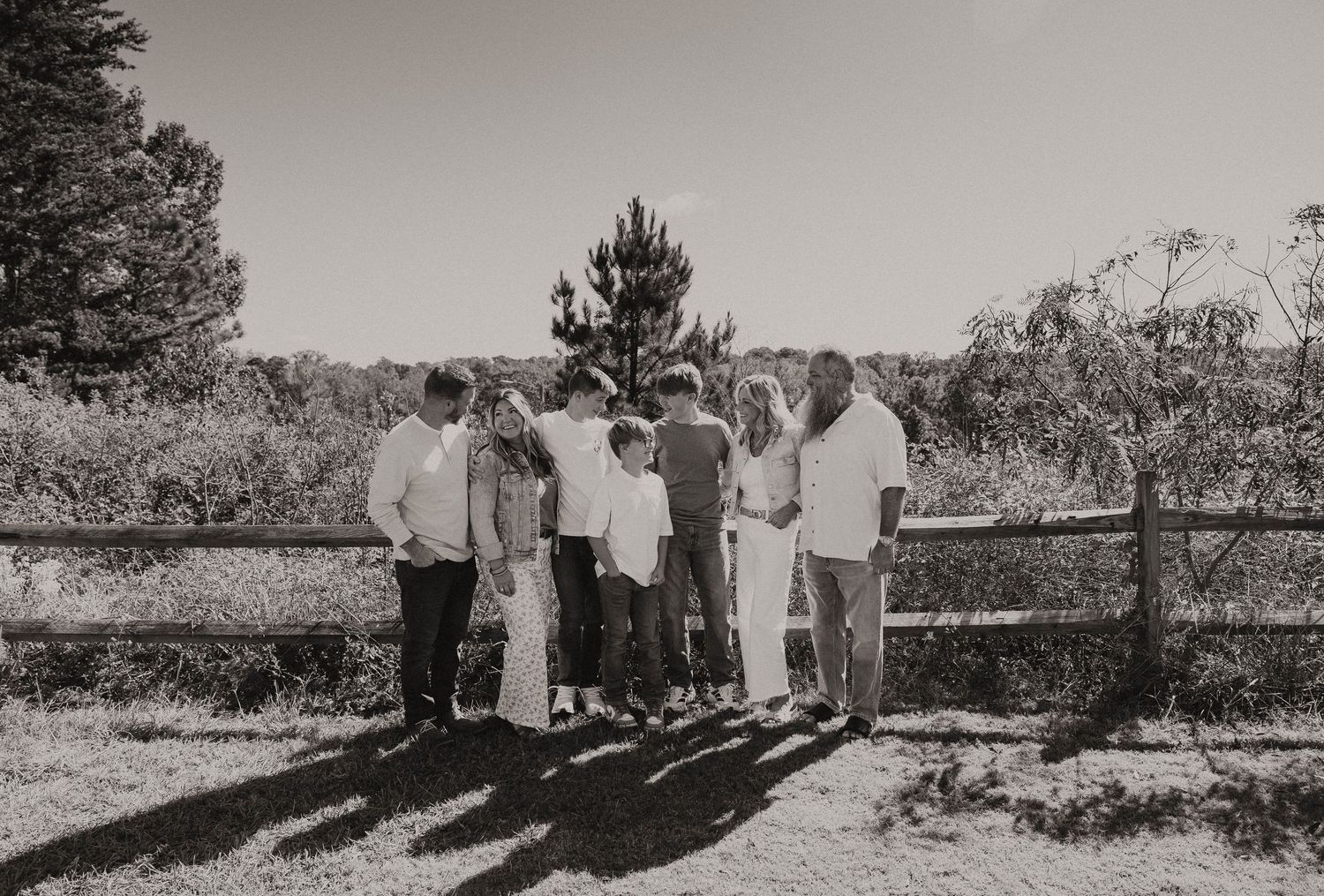 Black and white photo of large family group standing together by wooden fence in rural landscape.