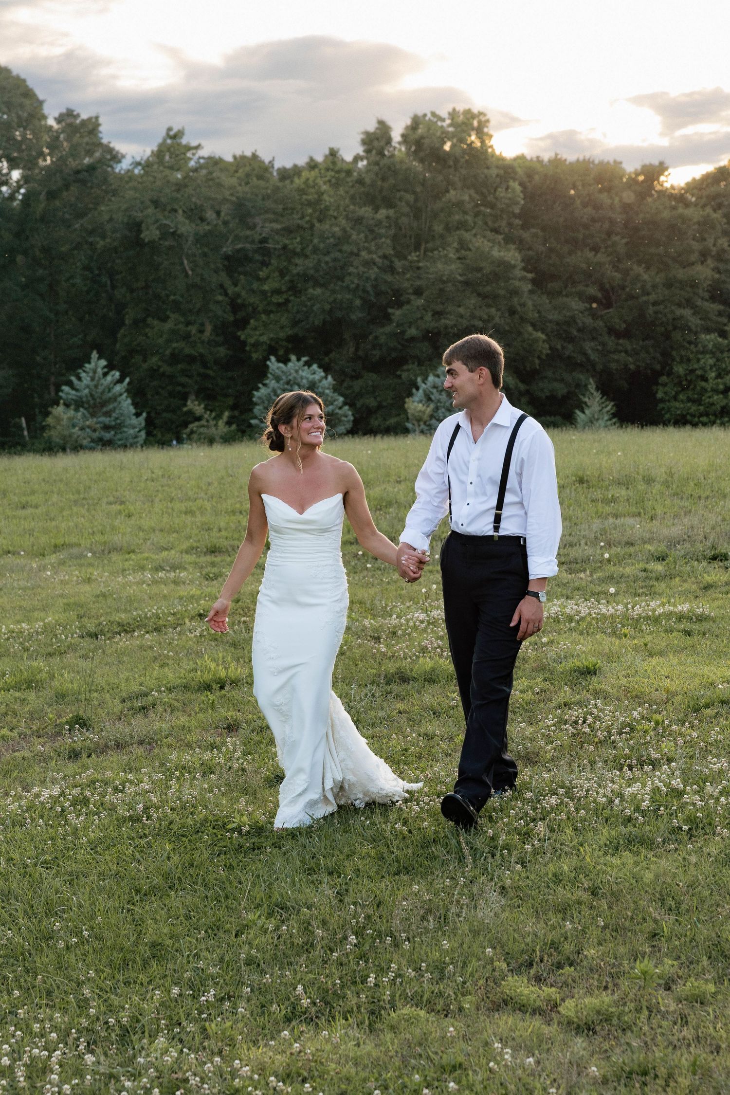 A bride and groom walk hand in hand through a sunlit green field at golden hour after their wedding.