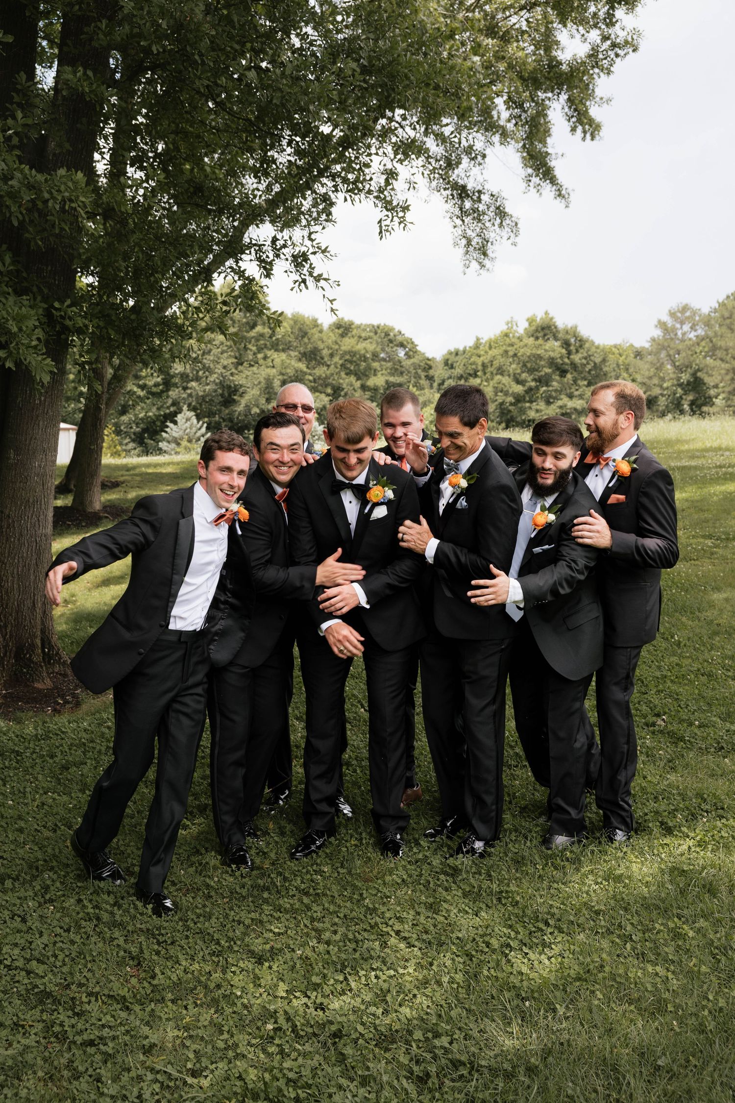 Groomsmen in black suits with orange boutonnieres laughing and posing together outdoors on green grass near trees.